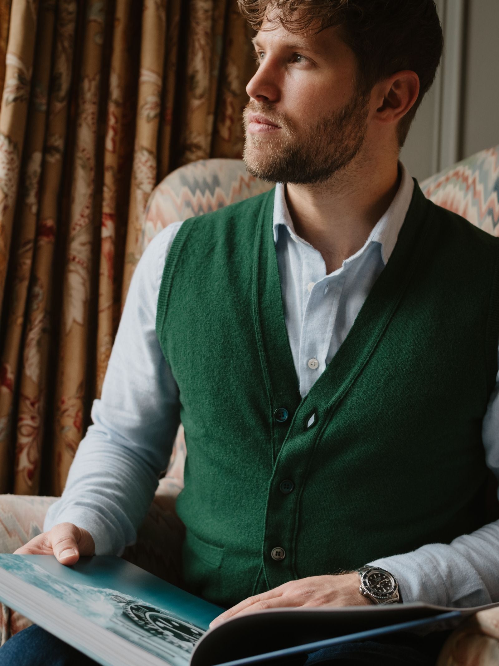 A man with a beard, in a light blue shirt and the Auld Stock Geelong Button Waistcoat by Campbell's of Beauly, sits in an armchair by patterned curtains holding an open book—an elegant addition to any country wardrobe.