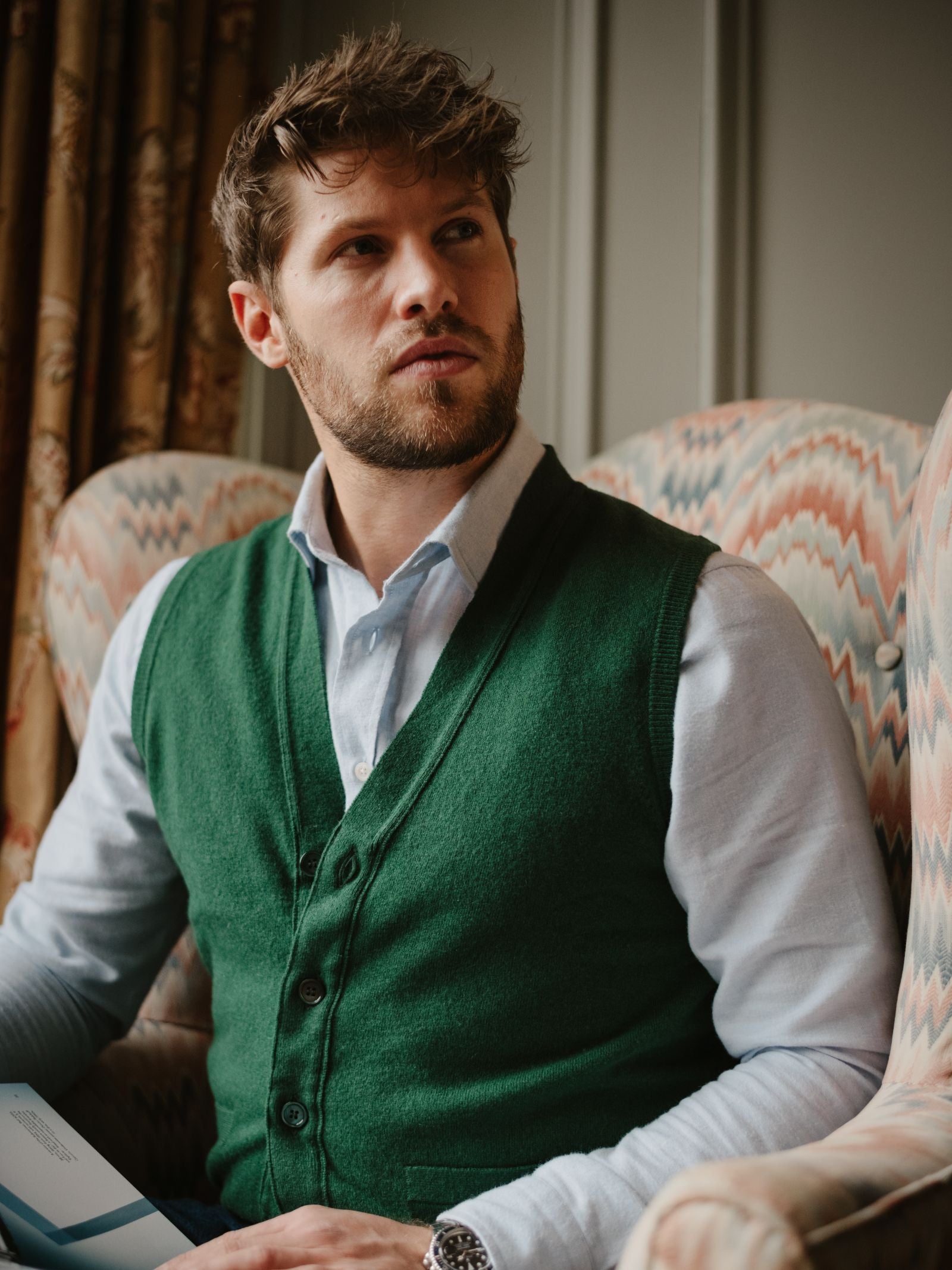 A man with short brown hair and a beard sits in an upholstered armchair, wearing a light blue shirt and the Campbell's of Beauly Auld Stock Geelong Button Waistcoat, made from merino wool, holding a book and looking thoughtfully to the side.