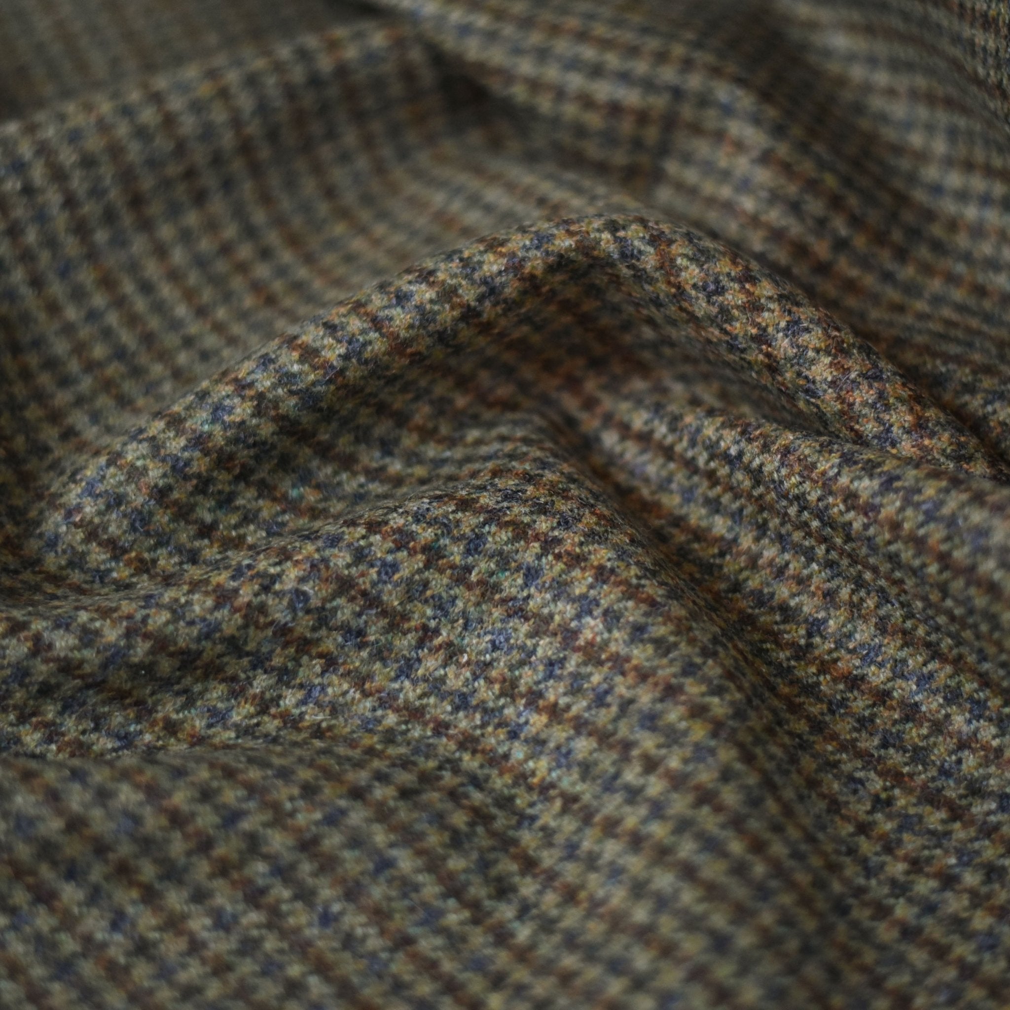 A close-up of House Tweed 84927 by Campbell's of Beauly features a brown and green houndstooth pattern, with gentle folds and shadows emphasizing the textured fabric’s Scottish tweed heritage.