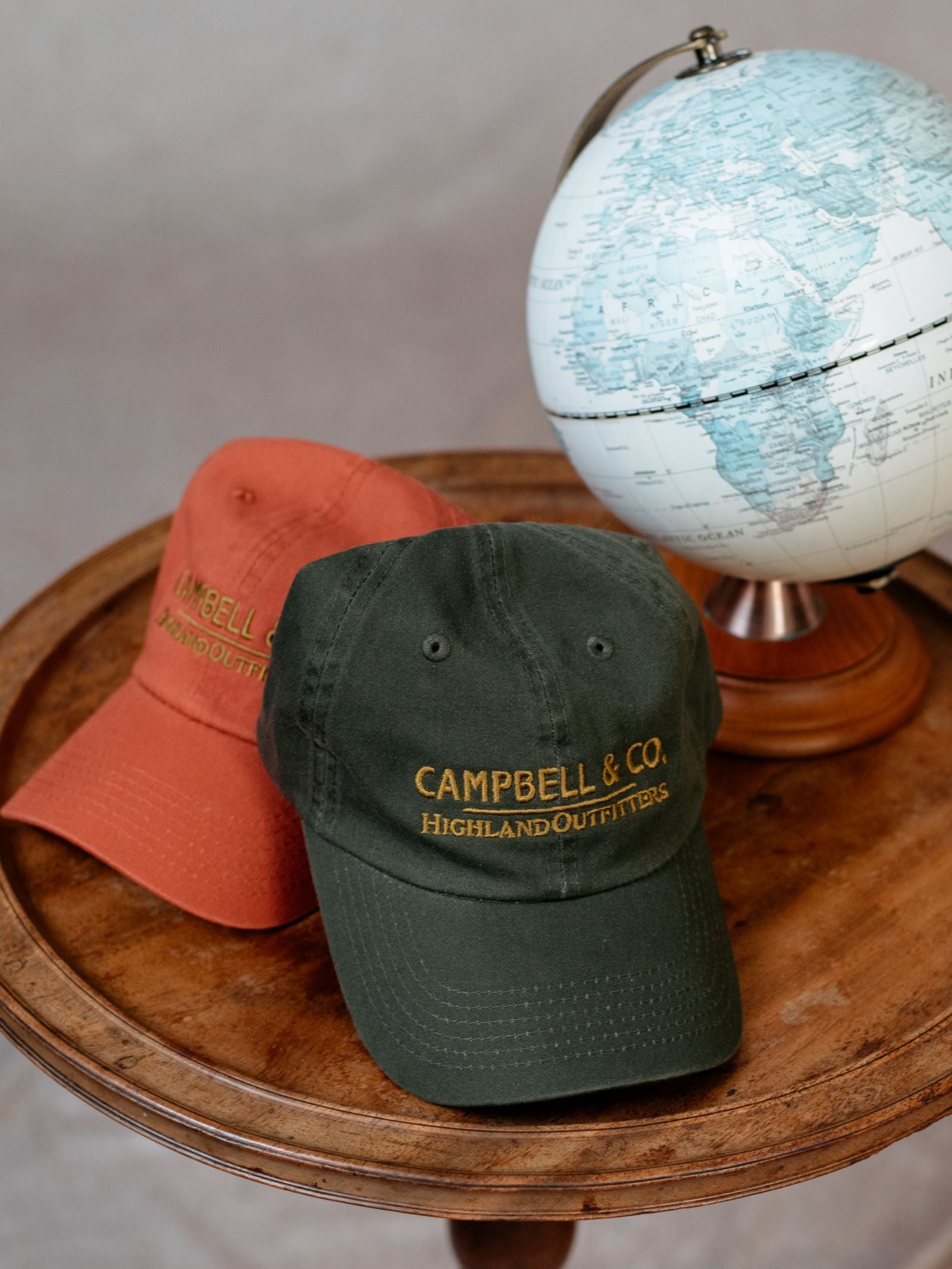 Two Unisex Campbell's Baseball Caps by Campbell's of Beauly—one dark green, one rust orange—featuring "CAMPBELL & CO. HIGHLAND OUTFITTERS," sit on a round wooden table beside a blue and white globe; ideal for daily wear.