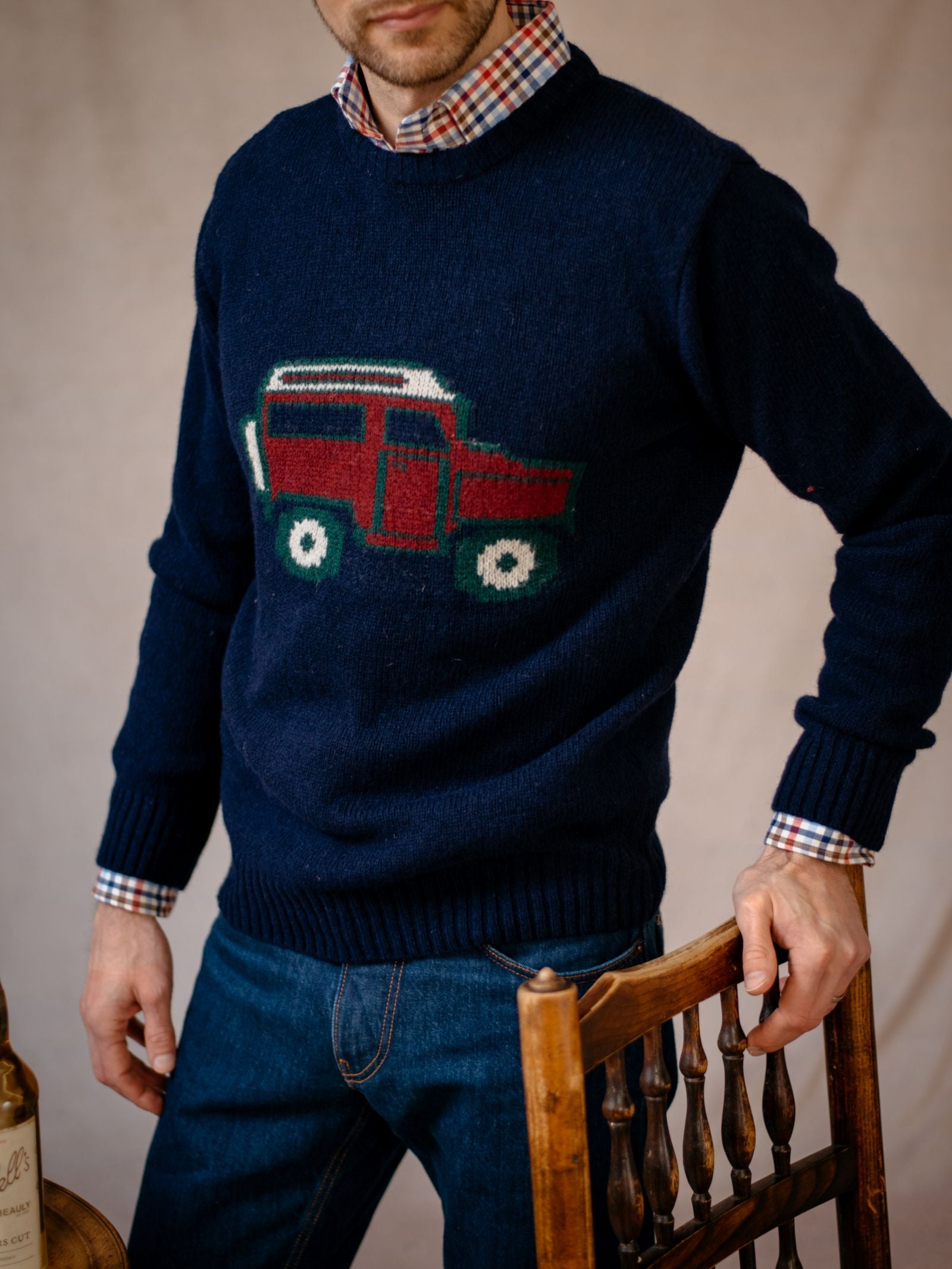 A man in a Campbell's of Beauly Unisex Land Rover Defender Jumper, navy with red and green car motif, over a plaid shirt and jeans stands by a wooden chair. His face is partially out of frame, evoking classic British motoring style.