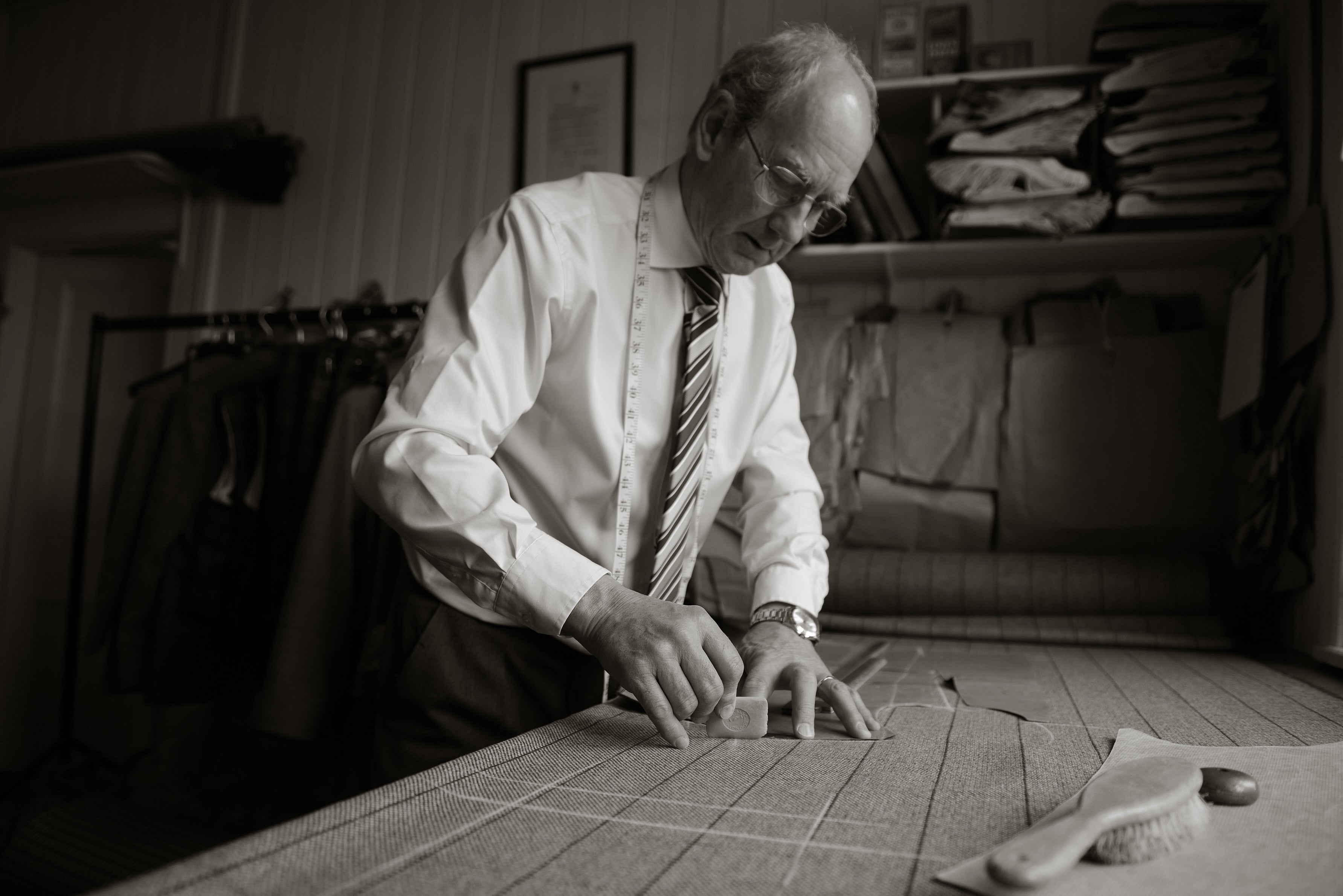 An older man in a shirt and tie measures fabric on a worktable in a tailor’s workshop, surrounded by fabric rolls, hanging clothes, and sewing tools.