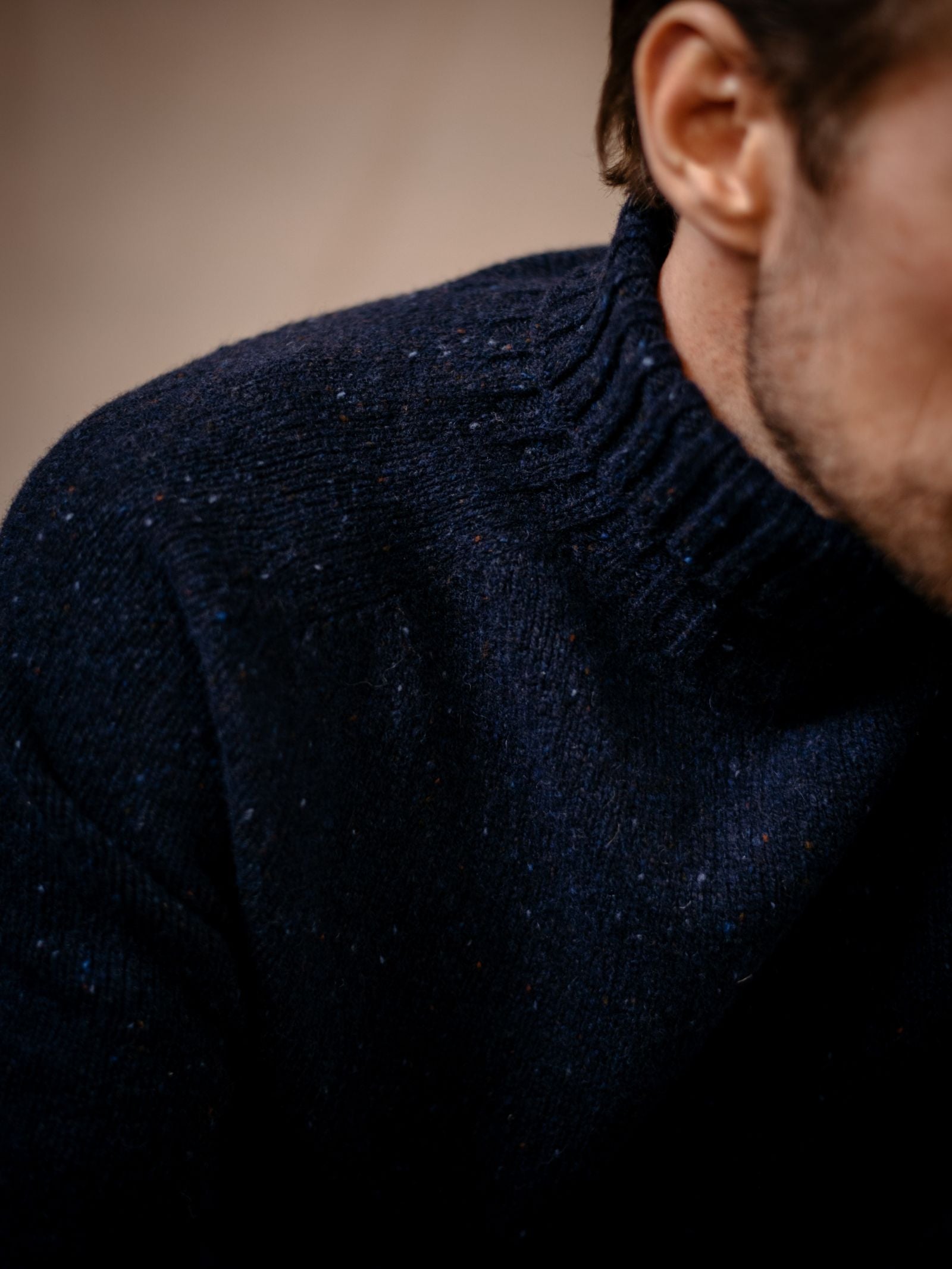 A close-up of a person in the Campbell's of Beauly Donegal Lambswool Roll Neck, a dark blue speckled sweater with a ribbed collar. The face is partially out of frame against a softly blurred background.
