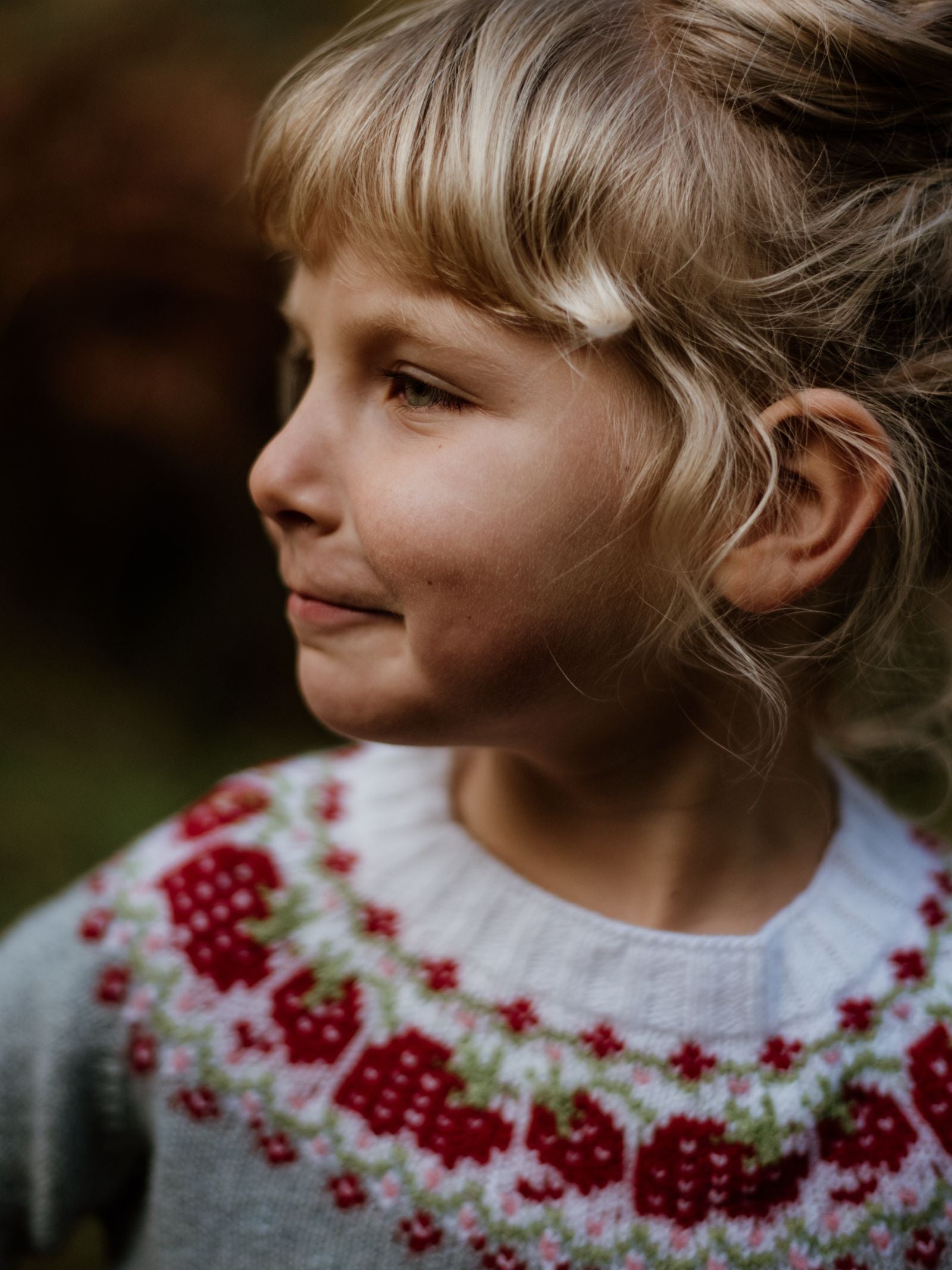 A young blonde child smiles while looking to the side, wearing the Campbells of Beauly Mini Strawberry Yoke Jumper, a light blue Geelong lambswool knit with a red, pink, and green Fairisle yoke, against a blurred outdoor background.