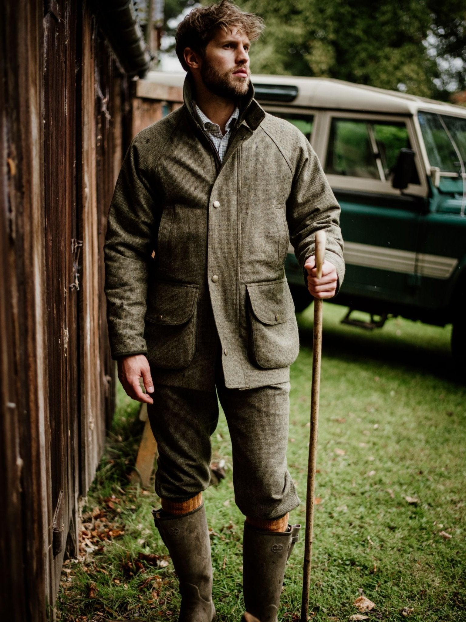 A man wearing Campbell's of Beauly Tweed Breeks and a Scottish tweed jacket, paired with tall rubber boots and a walking stick, stands by a wooden fence with a green classic SUV in the background on a grassy lawn.