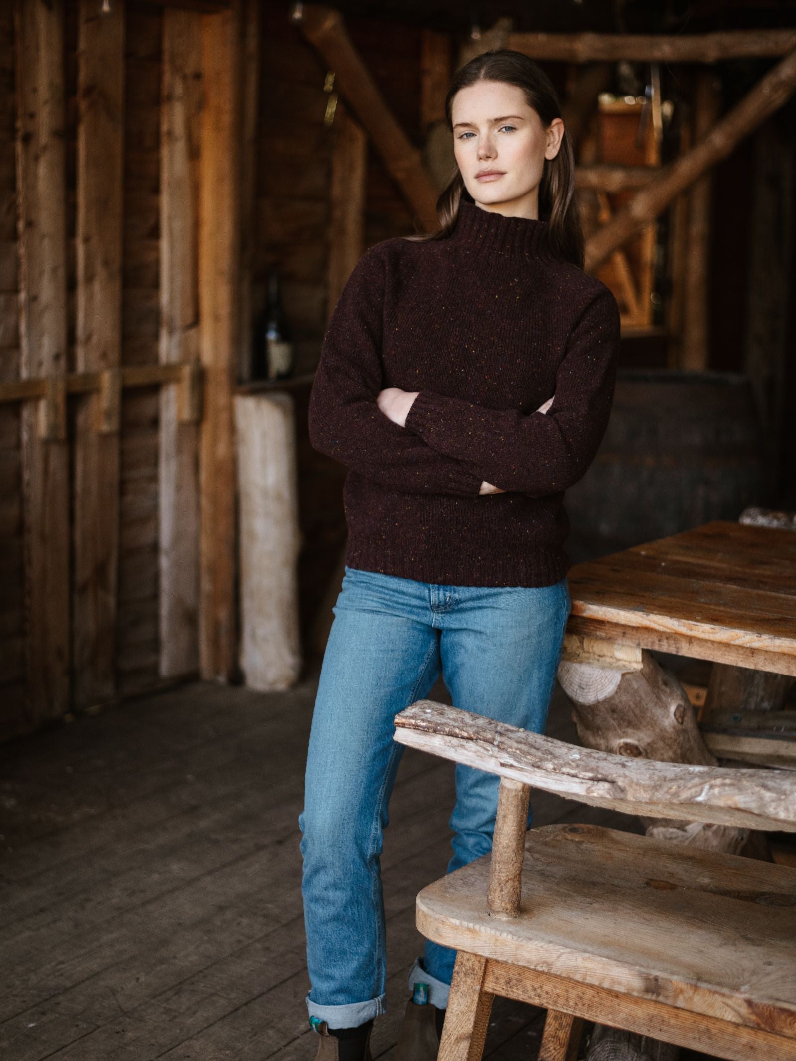 A woman in a dark Campbell's of Beauly Donegal Lambswool Funnel Neck Jumper and blue jeans stands indoors beside a rustic wooden table and benches, with a wooden wall and barrels in the background.