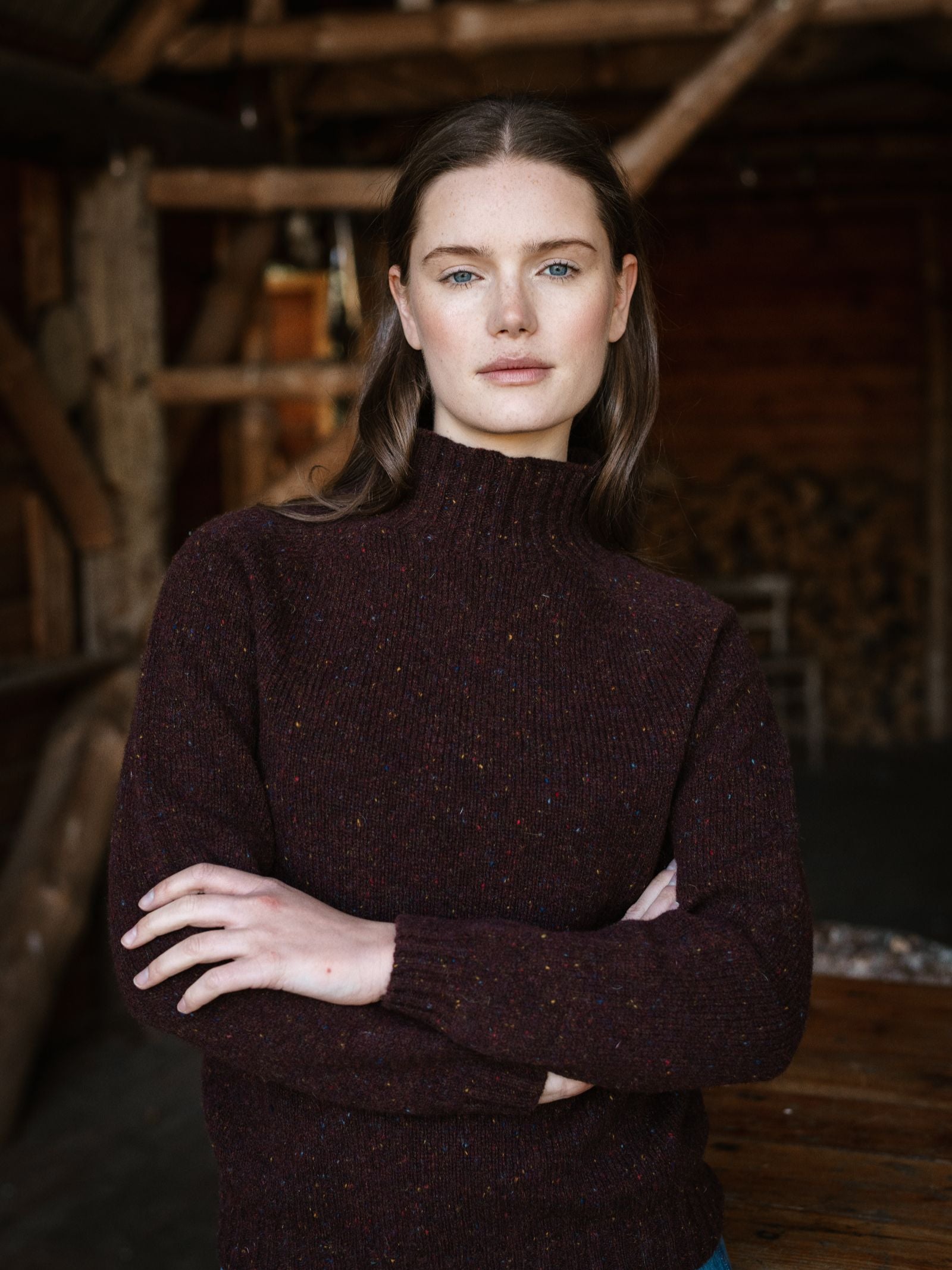A woman with long brown hair and blue eyes stands indoors with her arms crossed, wearing the Loganberry Donegal Lambswool Funnel Neck Jumper by Campbell's of Beauly, set against a rustic wooden, cabin-like background.