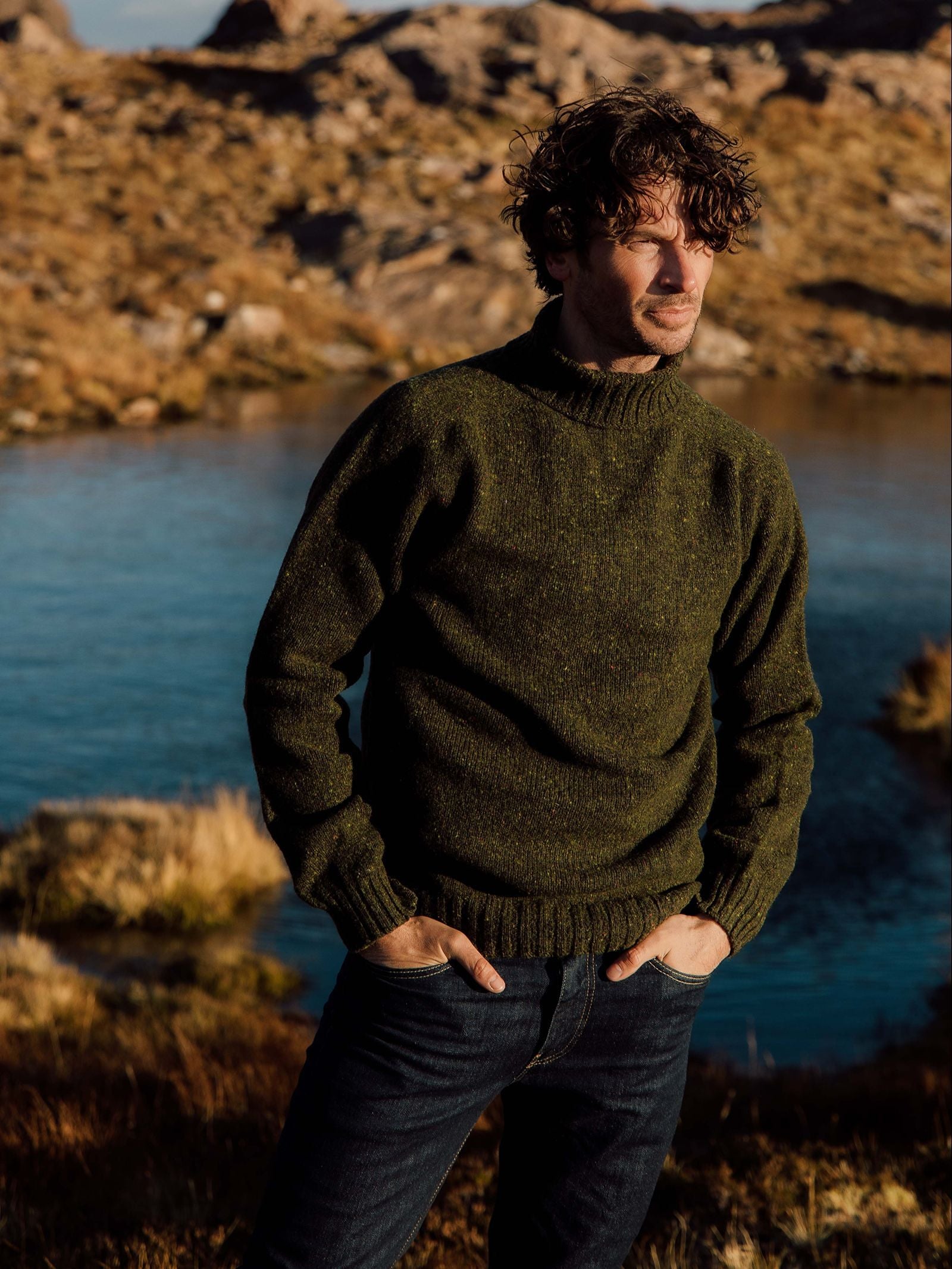 A man with wavy hair stands outdoors near a lake, surrounded by rocky hills and brown grass, wearing the Campbell’s of Beauly Donegal Lambswool Roll Neck sweater in dark green paired with jeans. This classic winter staple blends comfort and timeless style, making it ideal for chilly days.