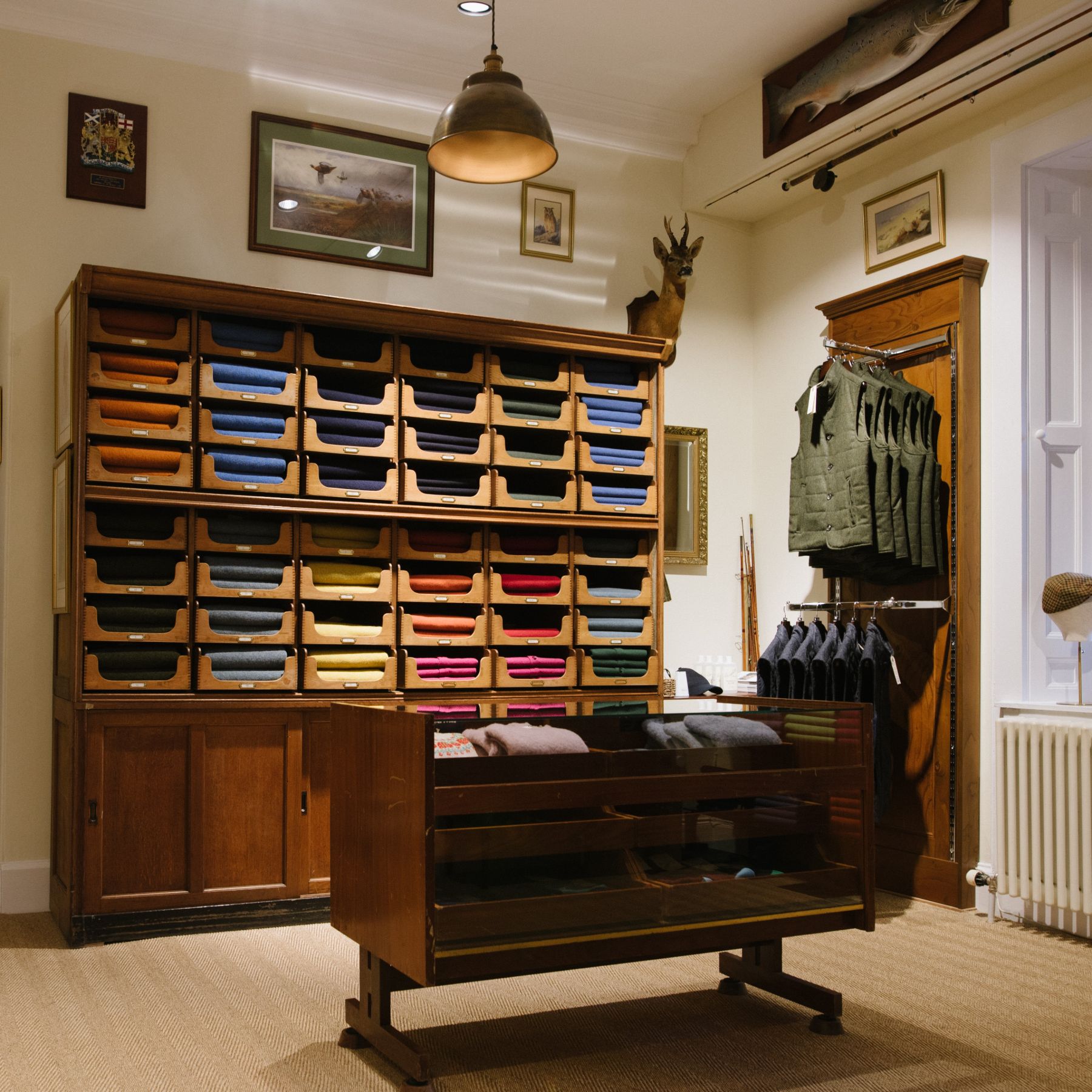 A well-organized room with wooden shelves holding neatly folded clothes in various colors, a glass display case, framed pictures on the walls, and several vests hanging on a wooden rack.