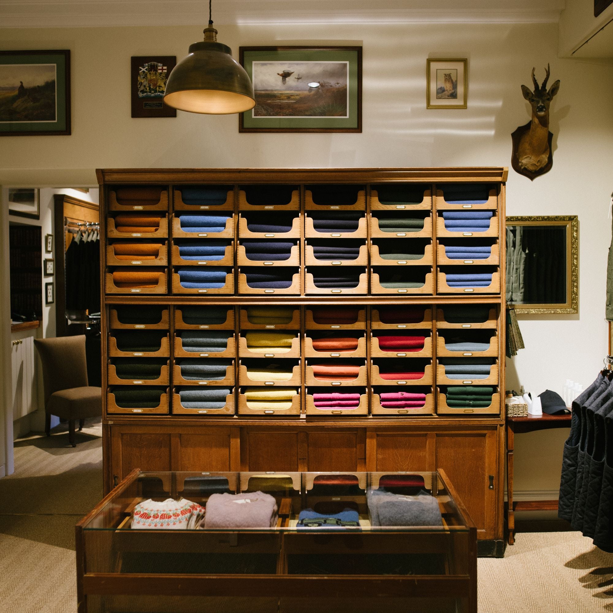 A wooden shelving unit holds neatly folded sweaters in various colors. A glass display case with more folded sweaters sits in front. The room has framed art, a hanging lamp, and a mounted deer head on the wall.