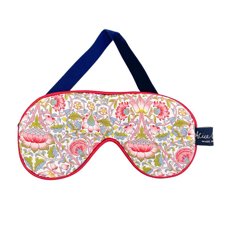 The Alice Caroline Liberty Print Eye Mask features a floral pattern in pink, green, and white, with a navy blue strap and red trim. This luxury sleep mask has a soft finish and is detailed with a small fabric tag.