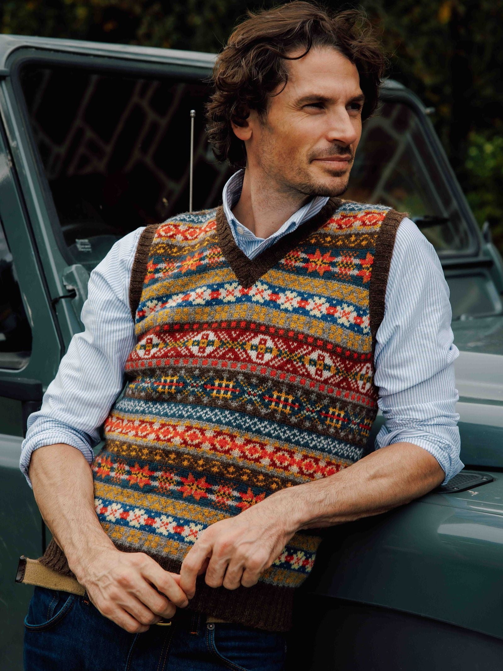 A man with wavy hair leans against a gray vehicle, wearing the Campbell’s of Beauly Fairisle Slipover—crafted from soft Shetland wool—layered over a light blue collared shirt and jeans, gazing thoughtfully to the side.