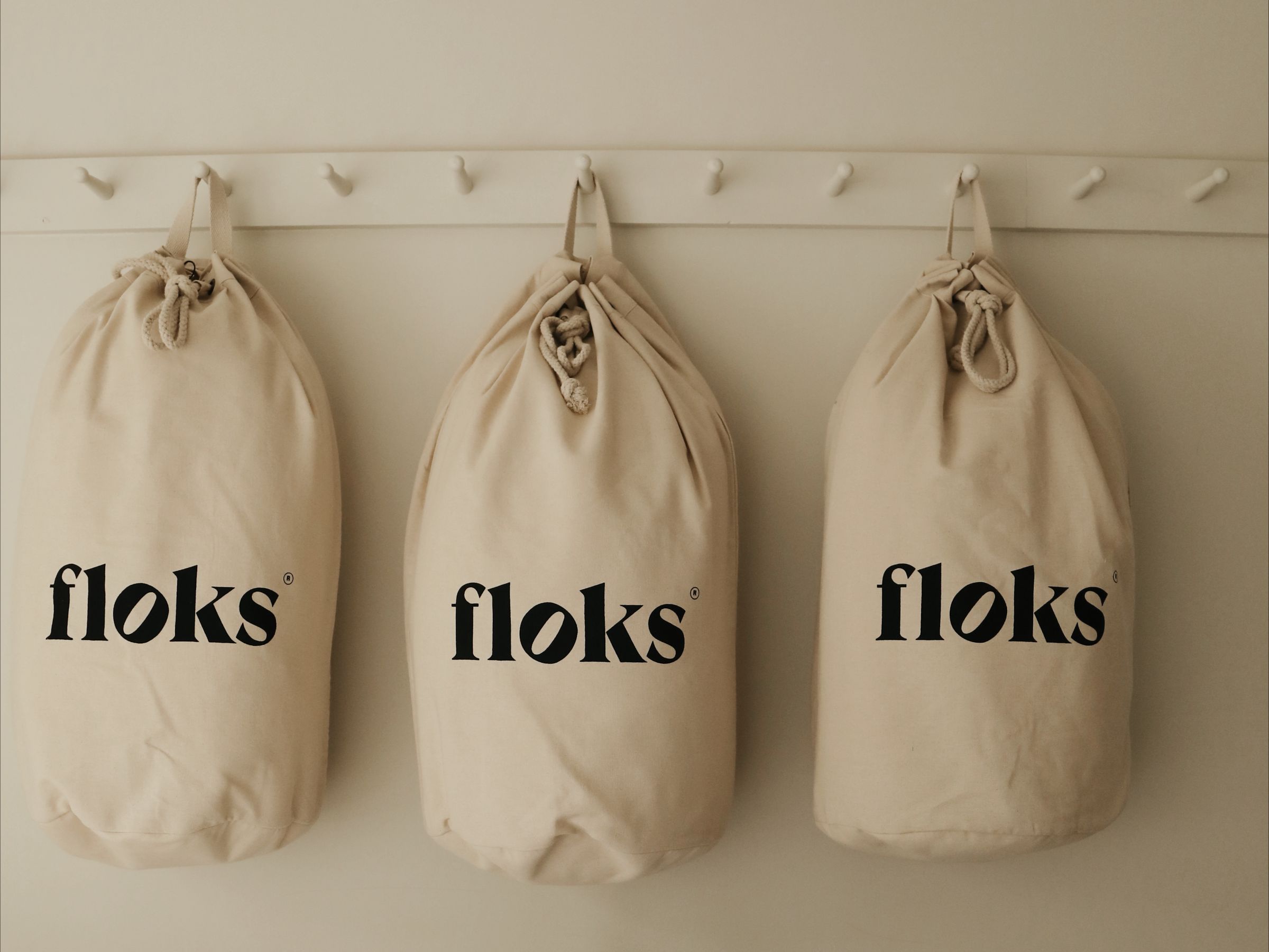 Three beige drawstring bags labeled floks hang side by side on white hooks against a light-colored wall.