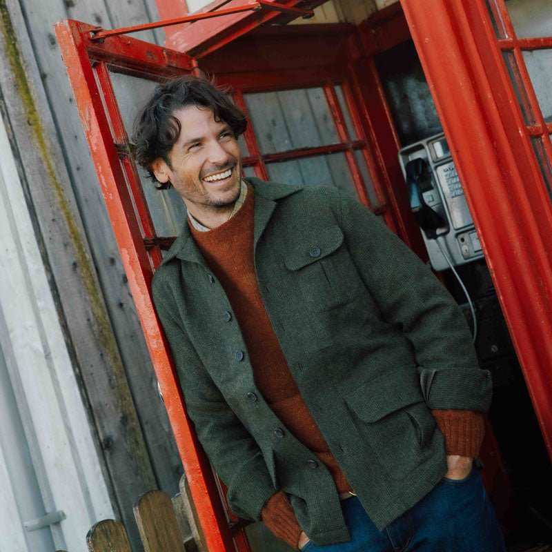 A man in the Campbell’s of Beauly Auld Stock Casual Shacket in Green Melange smiles while standing in the doorway of a red telephone booth, with a payphone visible behind him.