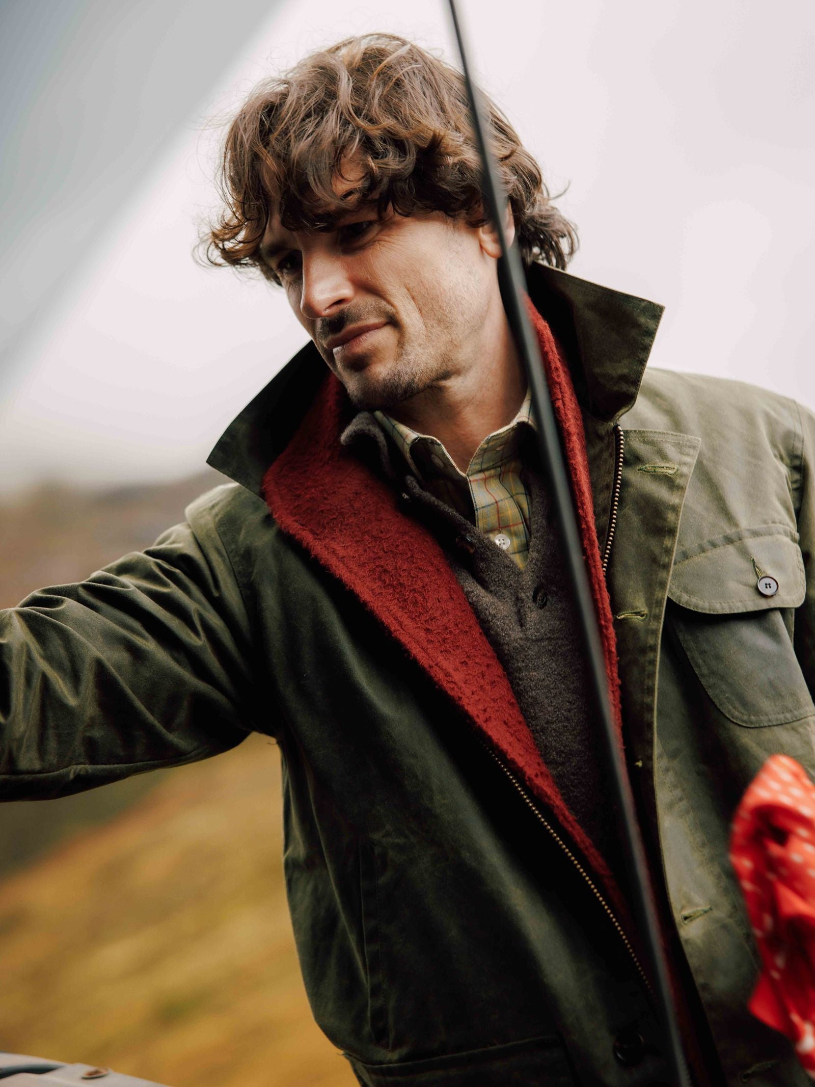 A man with wavy brown hair and a beard stands outdoors, looking down with a slight smile as he wears the Campbell’s of Beauly Garve Jacket—a green waxed cotton piece with a red lining that embodies effortless, field-ready style.