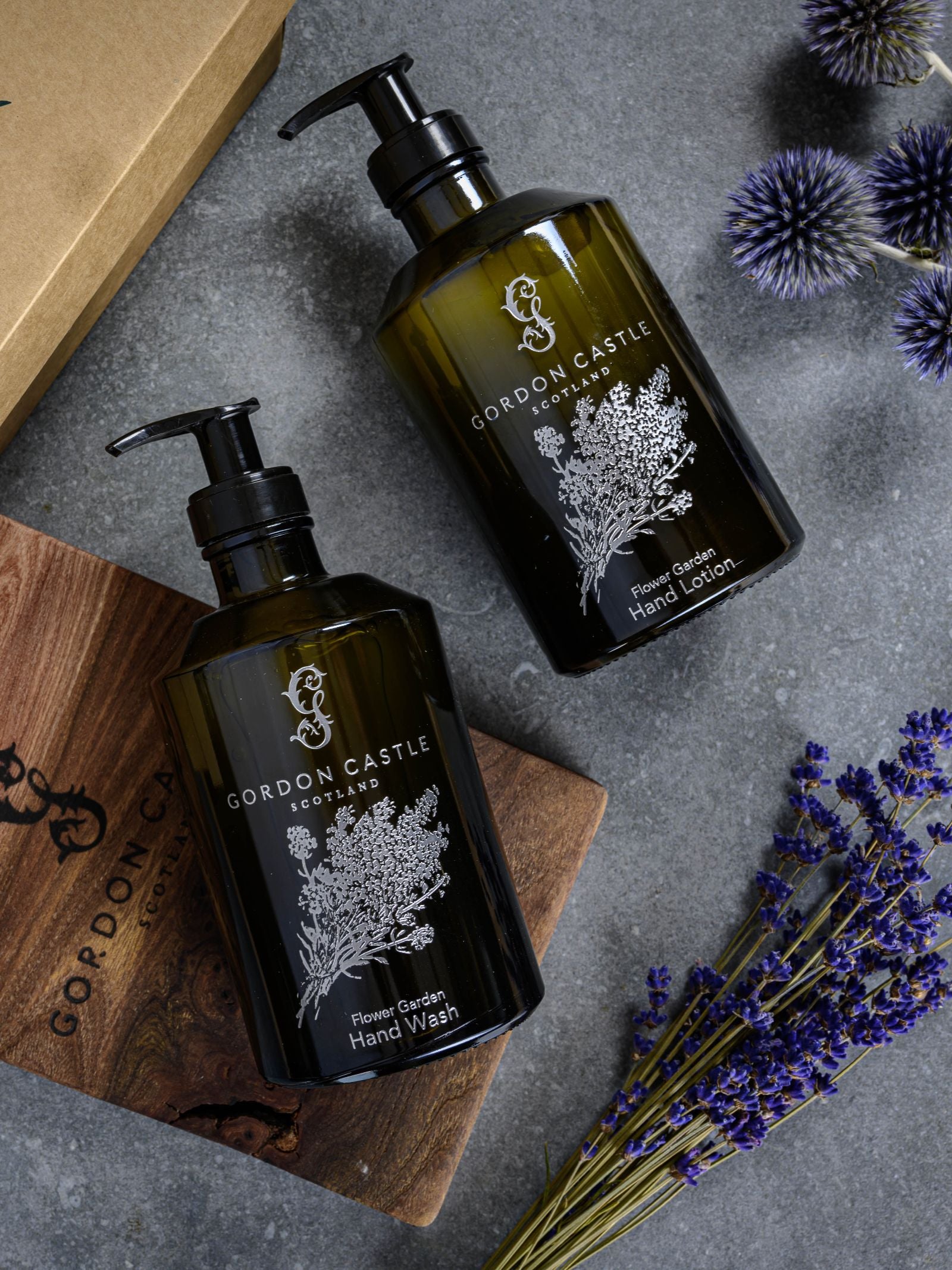 Two dark green refillable glass bottles of Gordon Castle's The Flower Garden Hand Lotion sit on a gray surface, surrounded by a wooden board, purple thistles, and lavender sprigs.