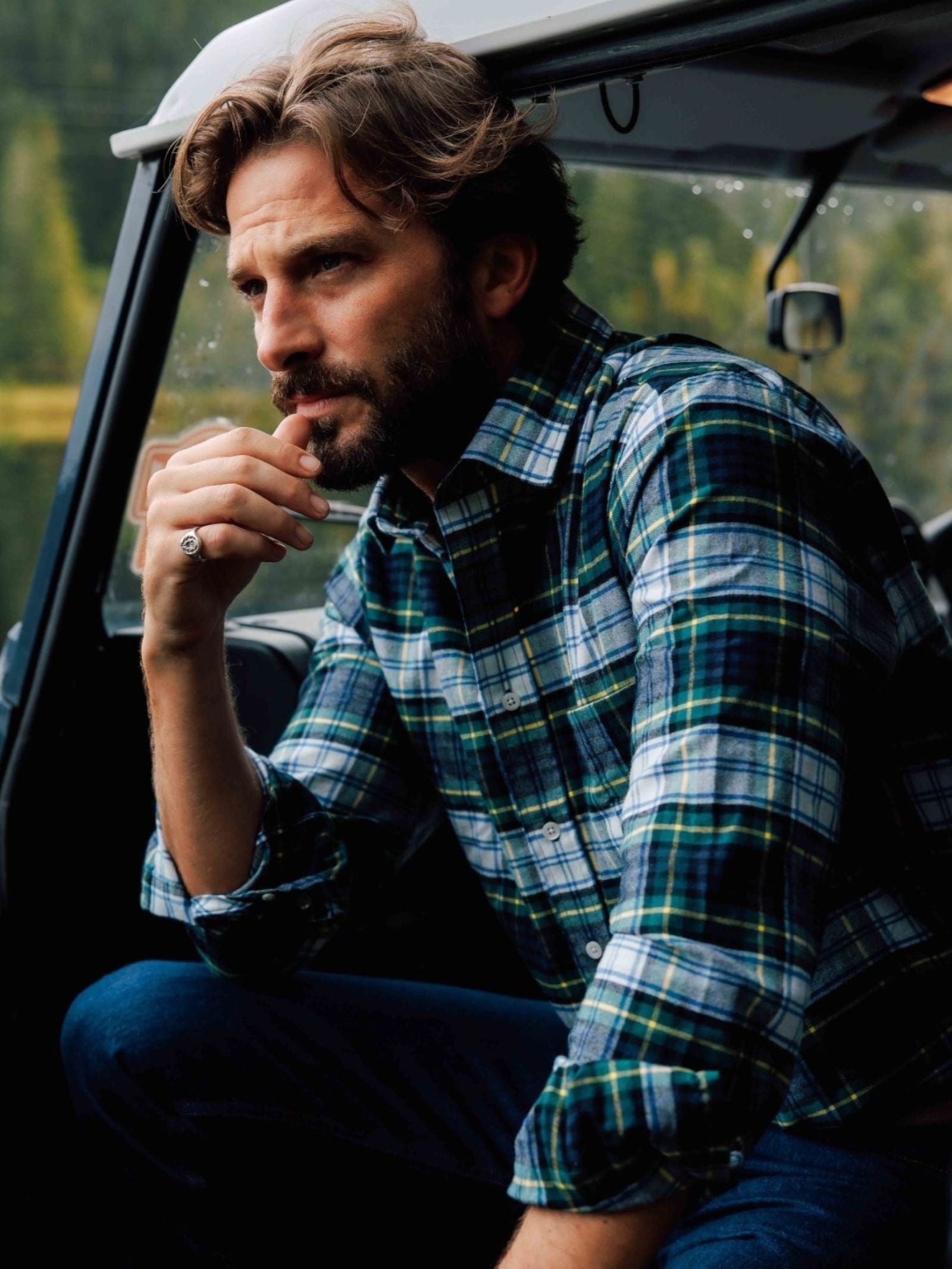 A man with wavy hair and a beard, wearing the Campbell's of Beauly Tartan Shirt and jeans, sits thoughtfully in a vehicle doorway with a forest in the background.