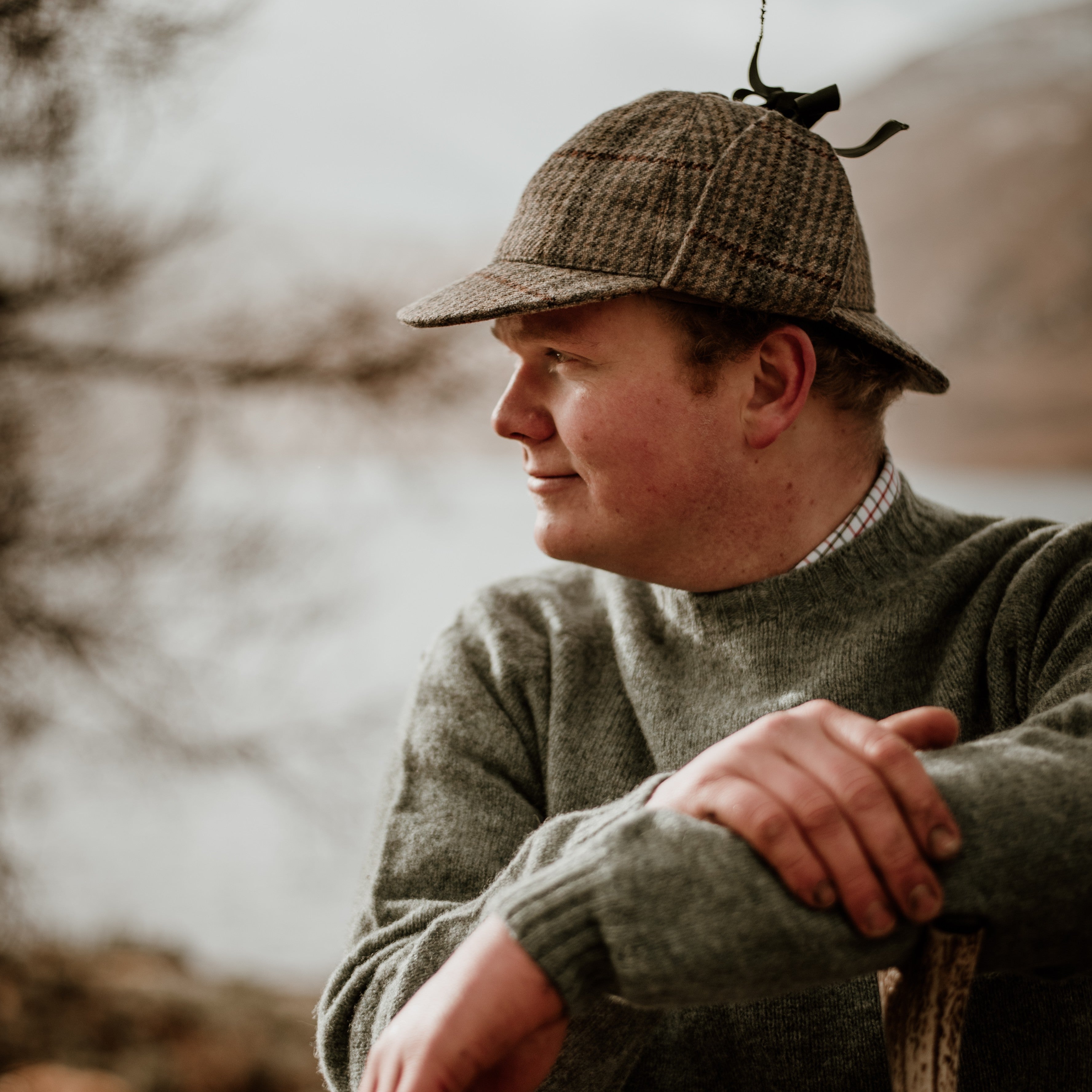 A man wearing a tweed deerstalker hat and gray sweater sits outdoors, looking to the side with folded hands resting on a chair, with a blurred natural landscape in the background.