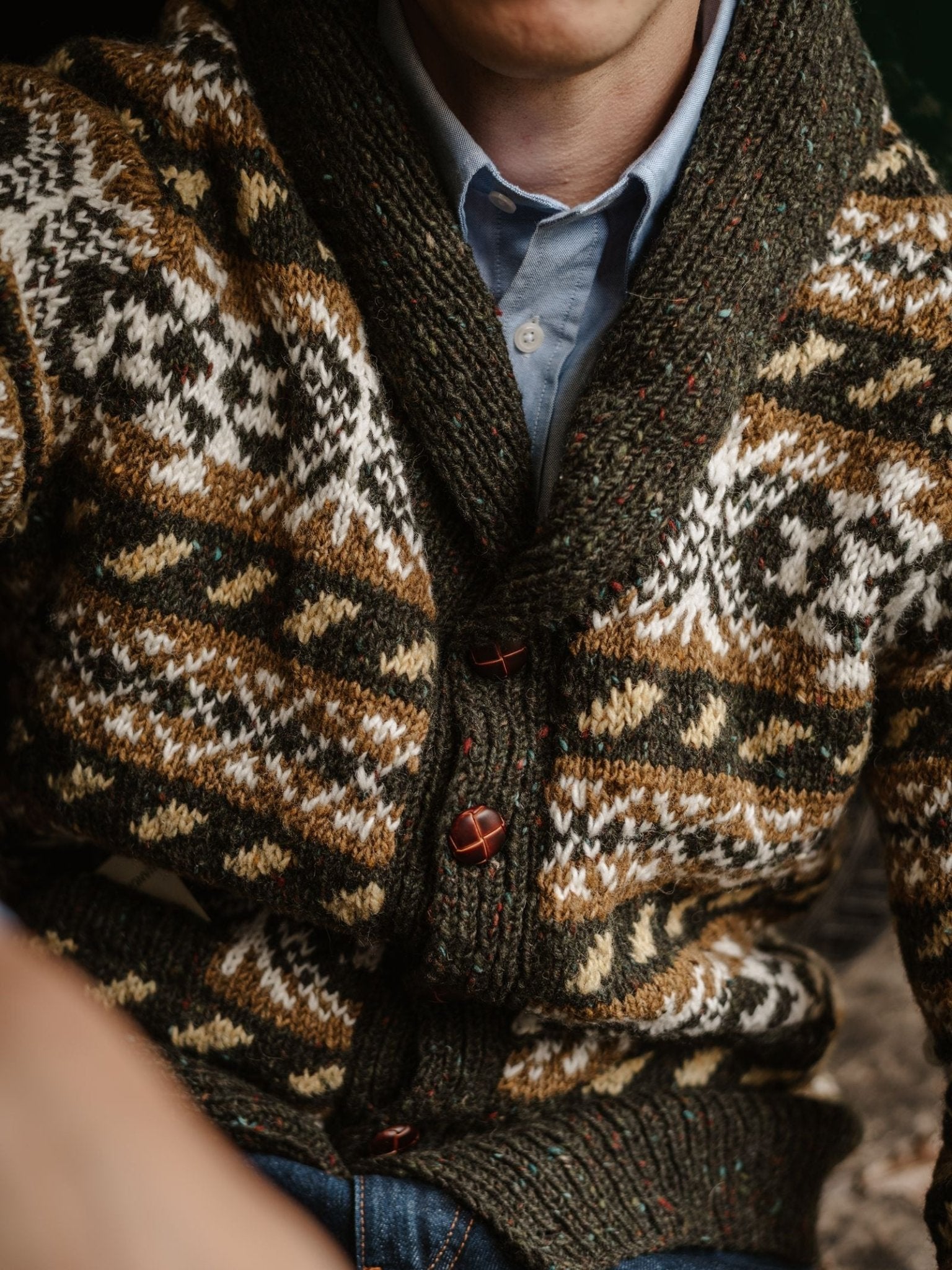 A person is wearing the Hand Knit Fairisle Shawl Cardigan by Campbell’s of Beauly—a chunky, patterned knit featuring brown and white designs and large buttons—layered over a blue shirt. The image is cropped, showing only part of their face and torso.