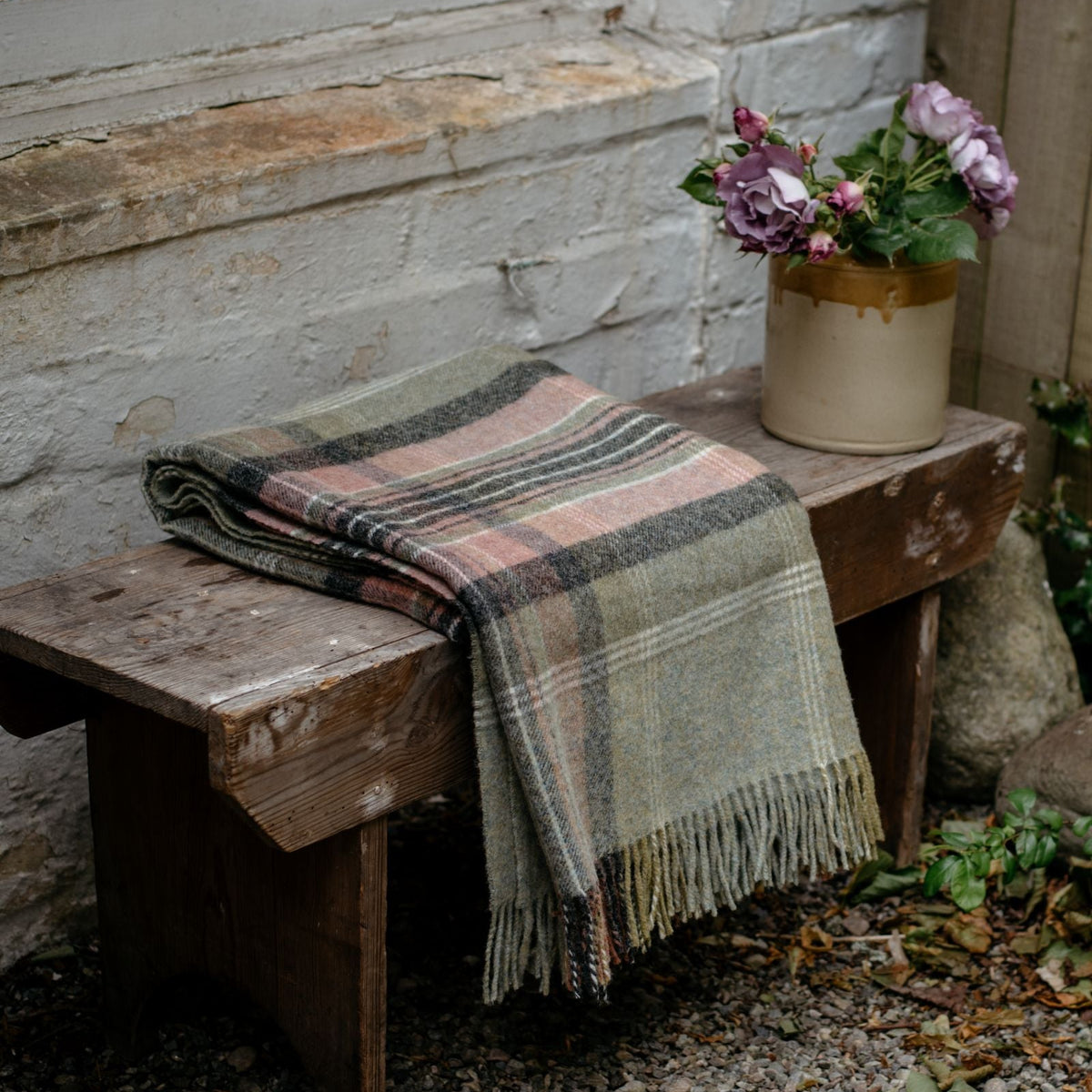 The Inchmore Lambswool Throw by Campbell’s of Beauly, folded with its fringe showing, adds Yorkshire charm on a rustic bench beside a mauve-flowered vase, set against a white brick wall and window.