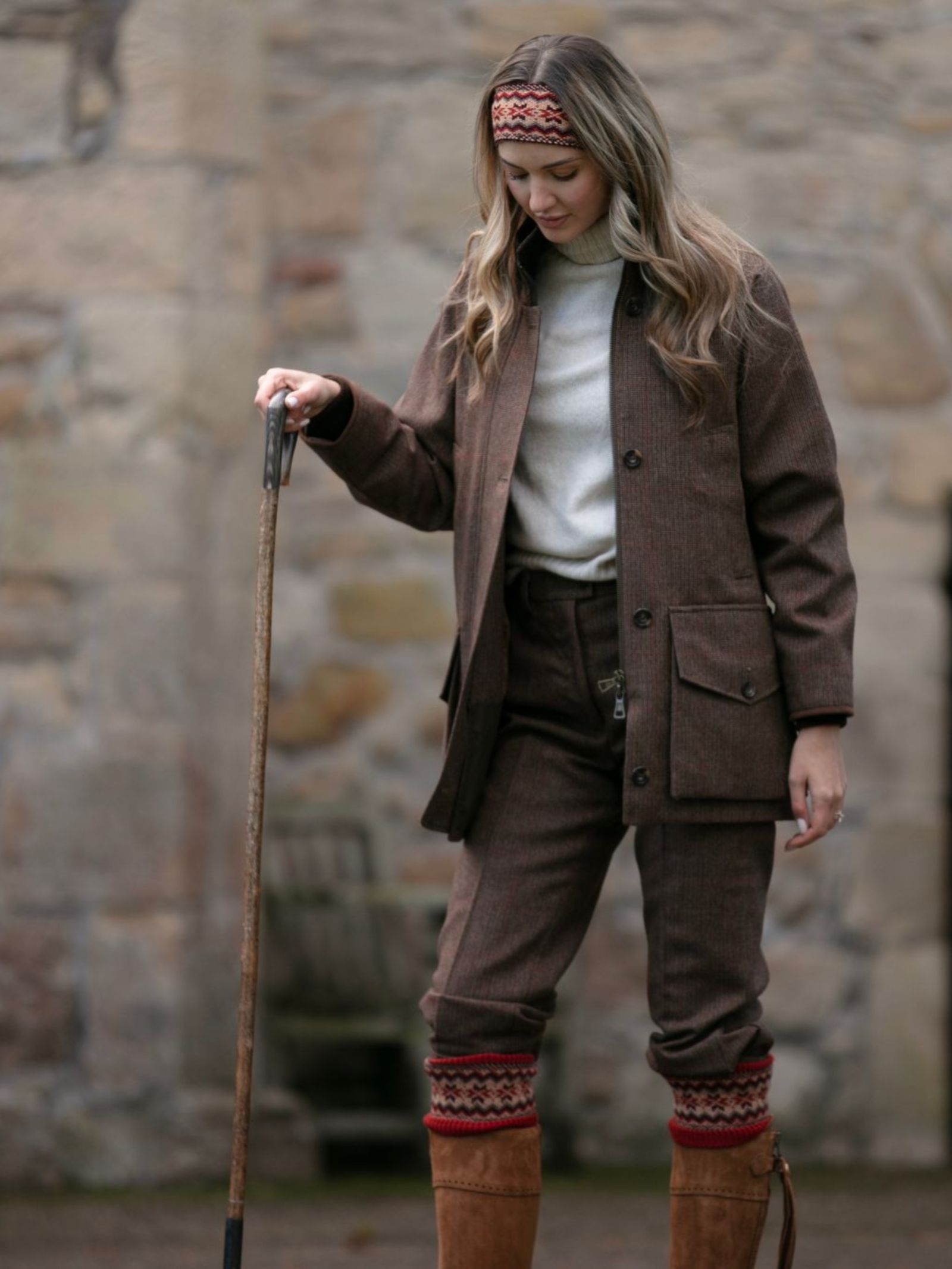 A woman wearing Campbell's of Beauly Fairisle Headband (100% wool) stands outdoors by a stone wall, dressed in a brown jacket, pants, and tan boots, holding a walking stick and evoking Scottish Borders charm.
