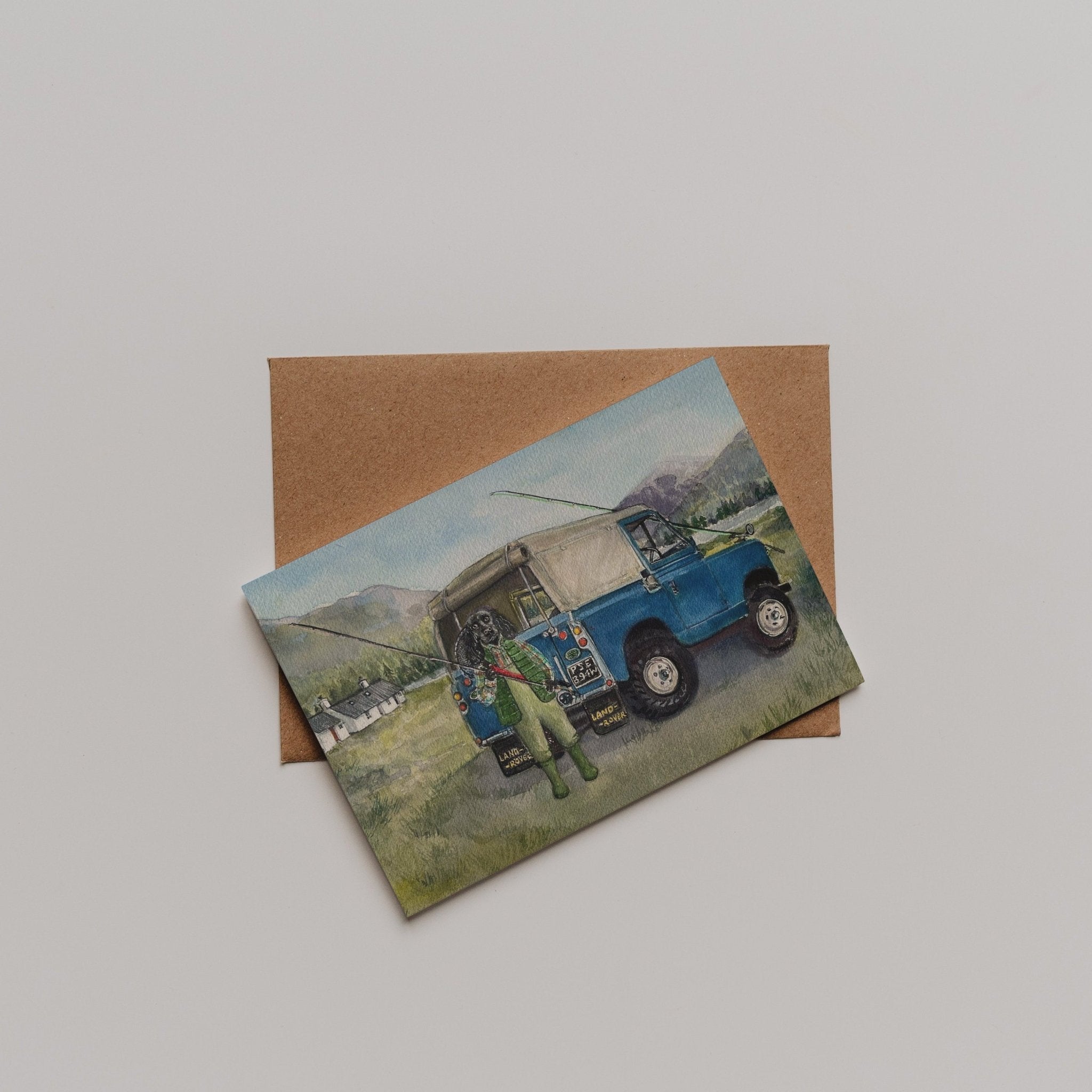 The Campbells of Beauly Land Rover Cocker Greeting Card features a Sophie Murray design: a blue vintage off-road vehicle on grass, mountains behind, with a person nearby. The card comes with a brown envelope.