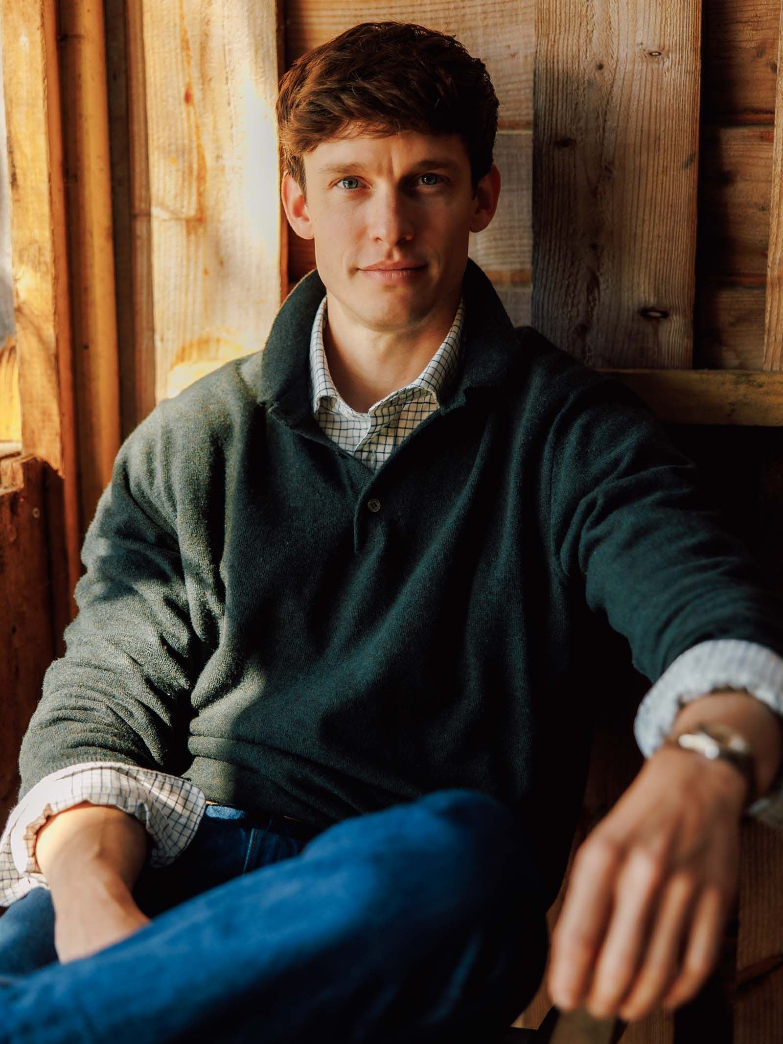 A man with short brown hair sits relaxed indoors, wearing the Campbell’s of Beauly Merino & Cashmere Rugby sweater layered over a checked shirt and paired with jeans. He looks at the camera with a slight smile, resting one arm on his knee against a wooden background.