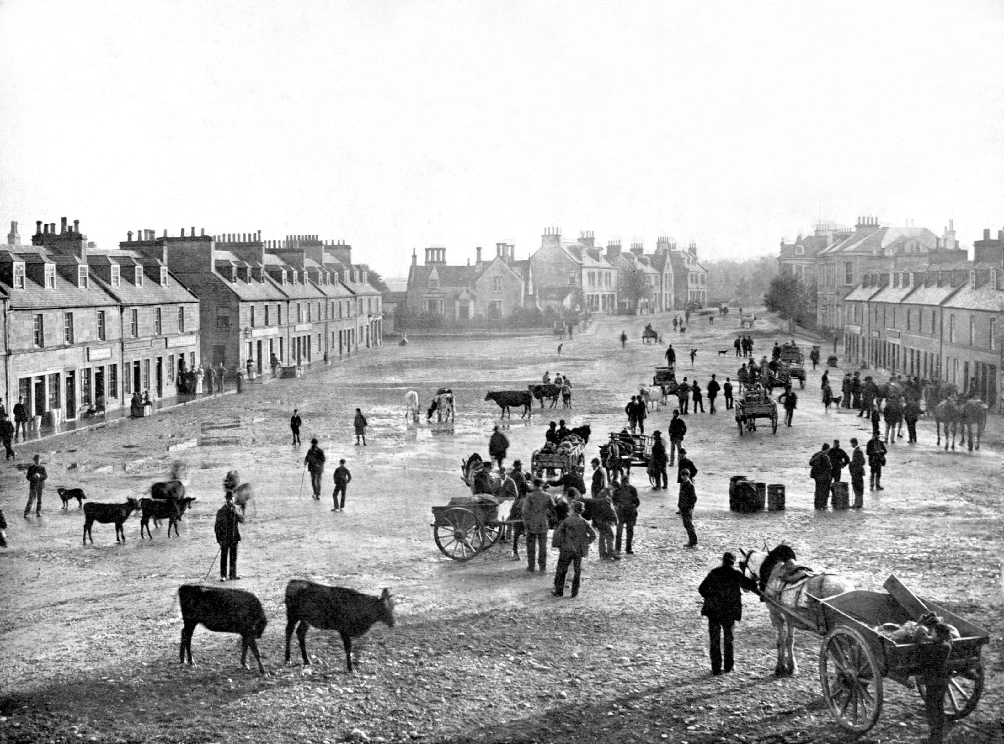 A black-and-white photo of a busy village street in the past, with people, horse-drawn carts, and cows scattered around open space, lined by rows of stone houses and shops on both sides.