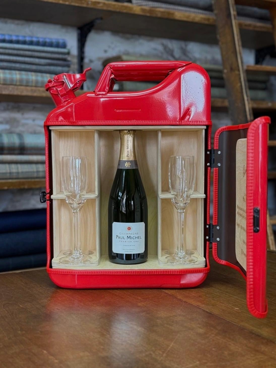 The Paul Michel Champagne Upcycled Mini Bar by Campbell's of Beauly is a red metal jerry can with a wooden interior, designed to hold a bottle of Paul Michel champagne and two flutes—perfect for impressing guests.