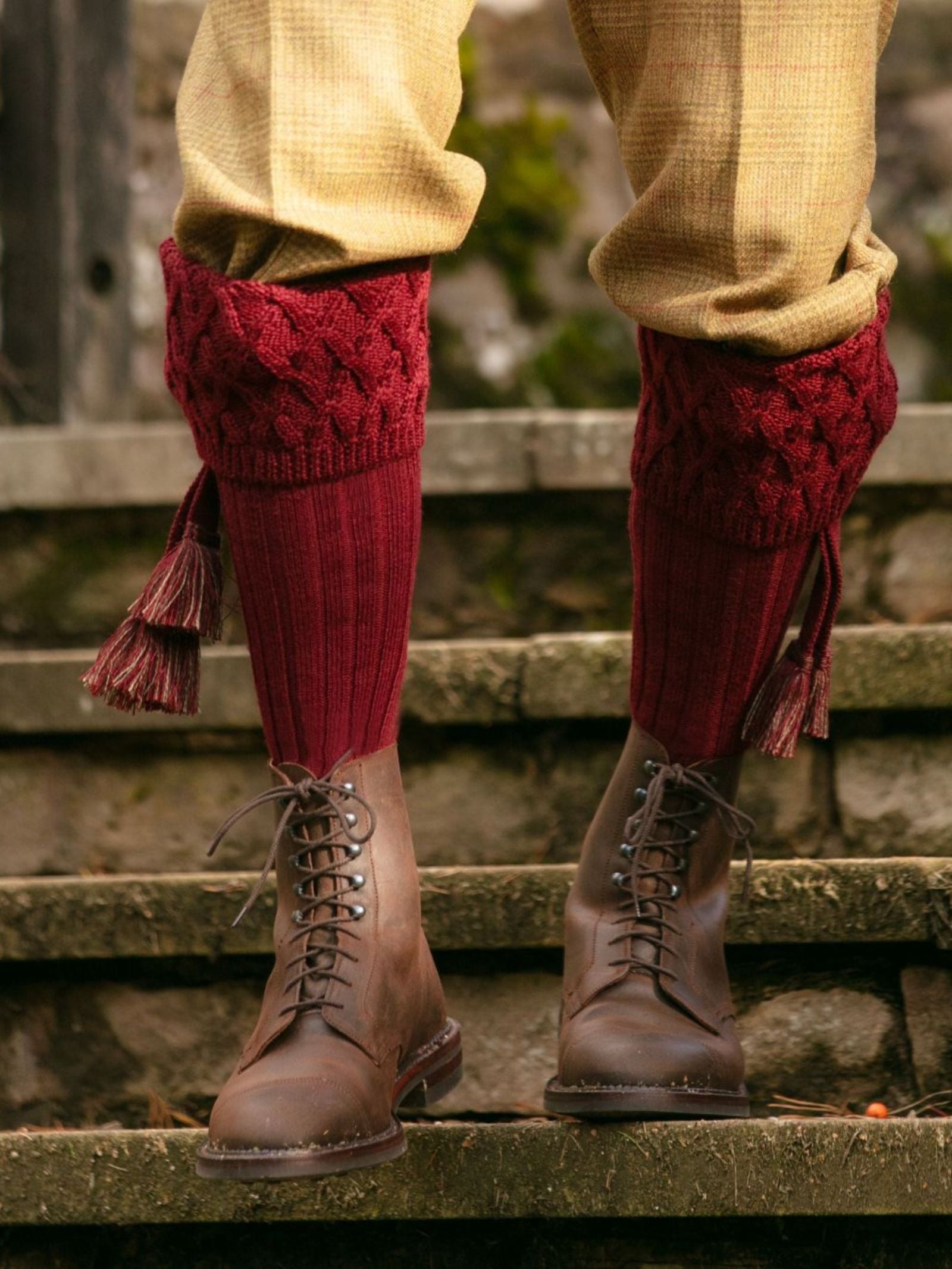A person sports tan trousers, brown lace-up boots, and Campbell’s of Beauly Merino Rannoch Shooting Socks in red with tassels while standing on stone steps outdoors for a classic country style look.