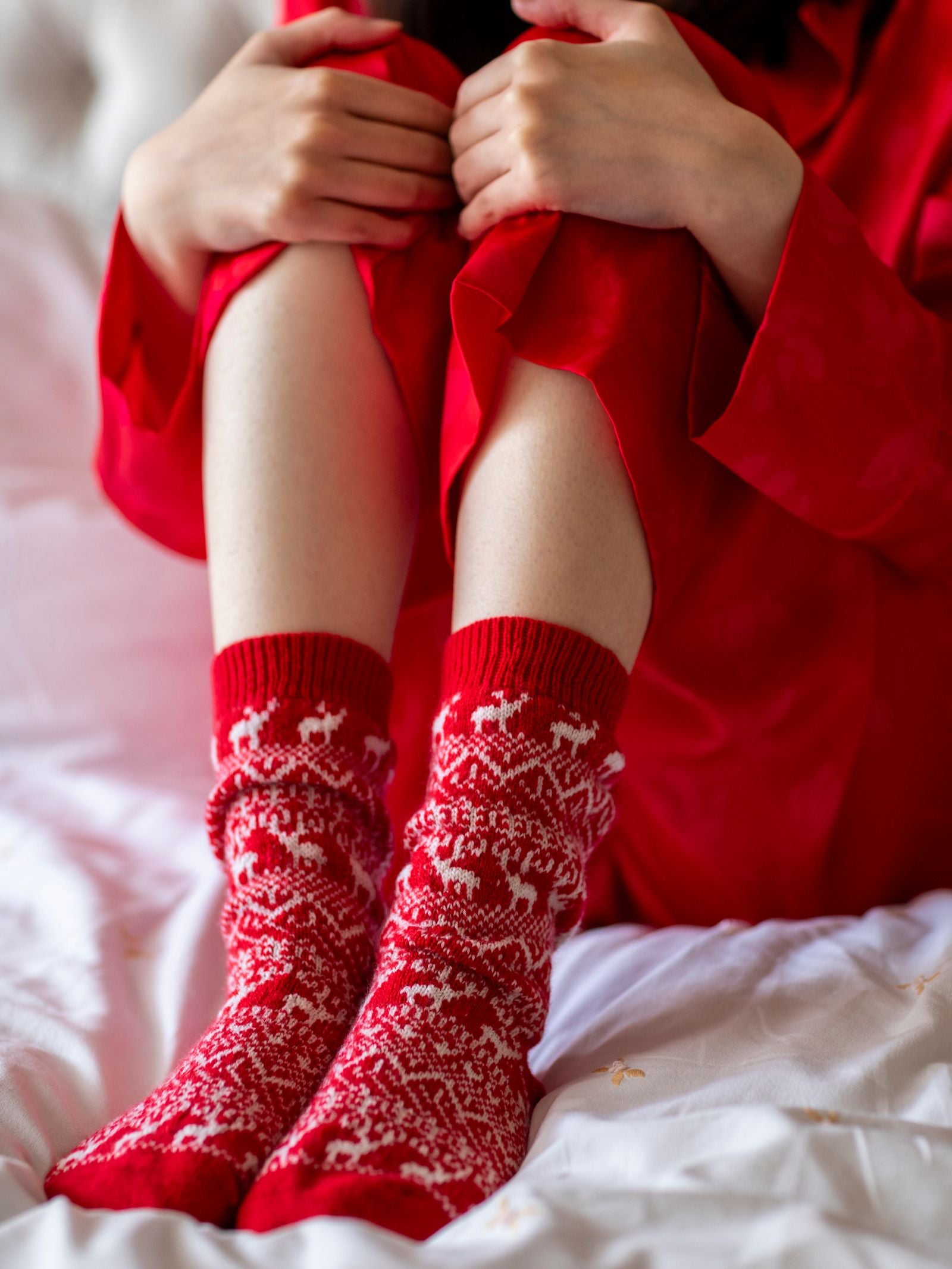 A person in red pajamas sits on a bed, hugging their knees and wearing Campbell’s of Beauly Cashmere Reindeer Socks in red.