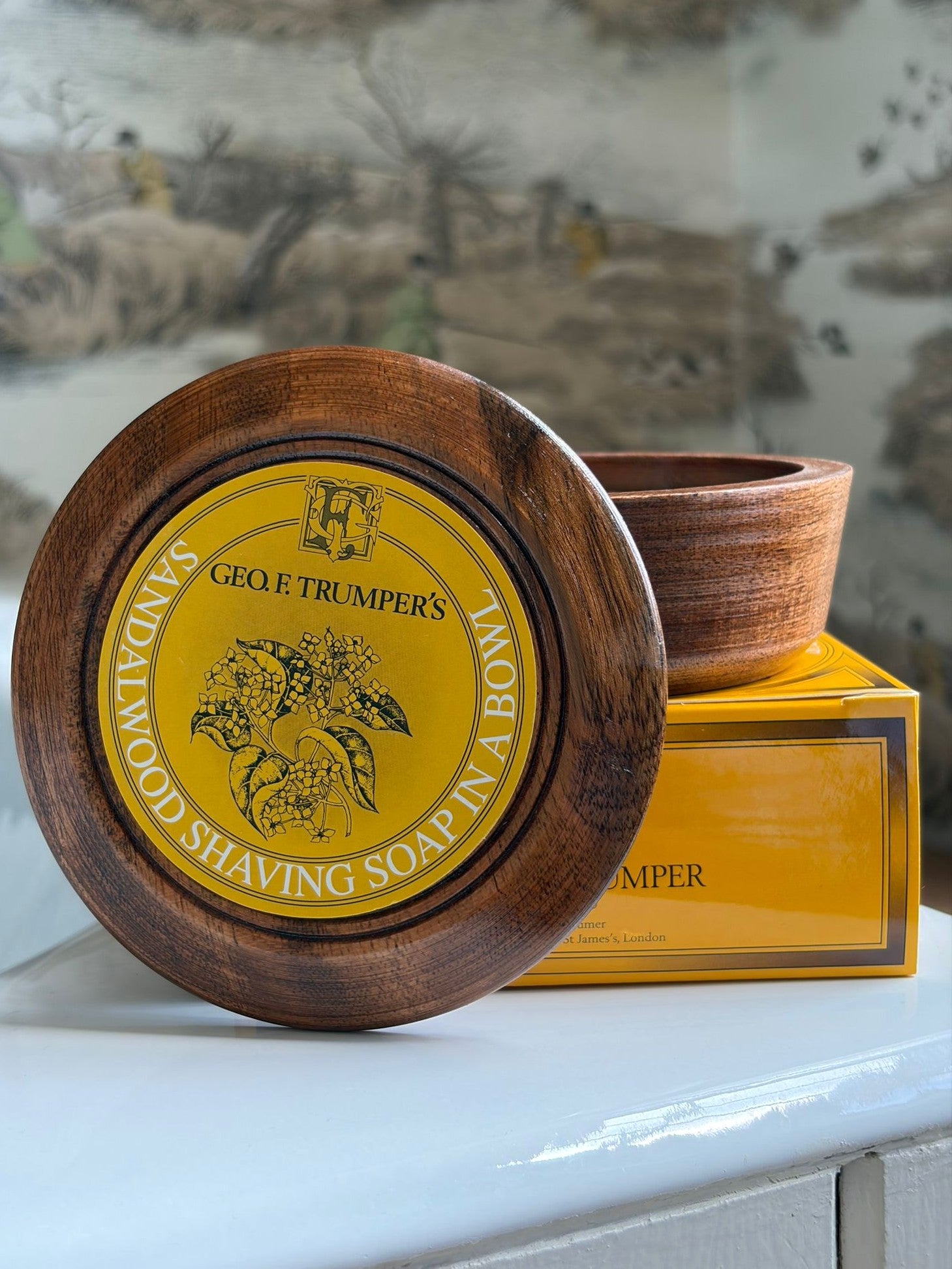 The Geo. F. Trumper Sandalwood Shaving Soap & Bowl, with its signature yellow label, rests on a matching box—an ideal choice for a luxurious wet shave, all set against a softly blurred scenic background.
