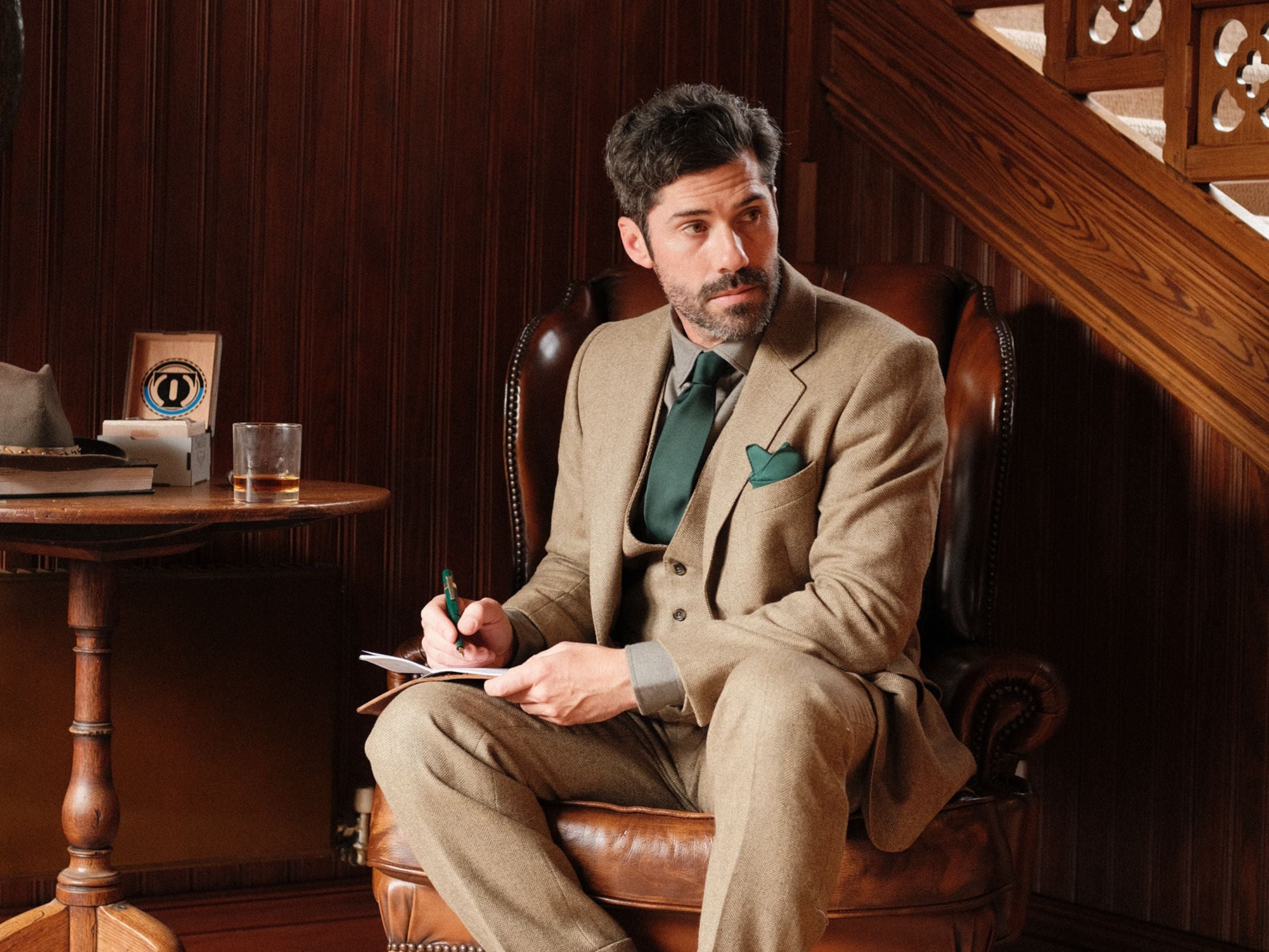 A man with dark hair and a beard, dressed in a tan suit with a green tie and pocket square, sits in a leather chair holding a pen and notepad. He looks to the side in a wood-paneled room near a staircase.