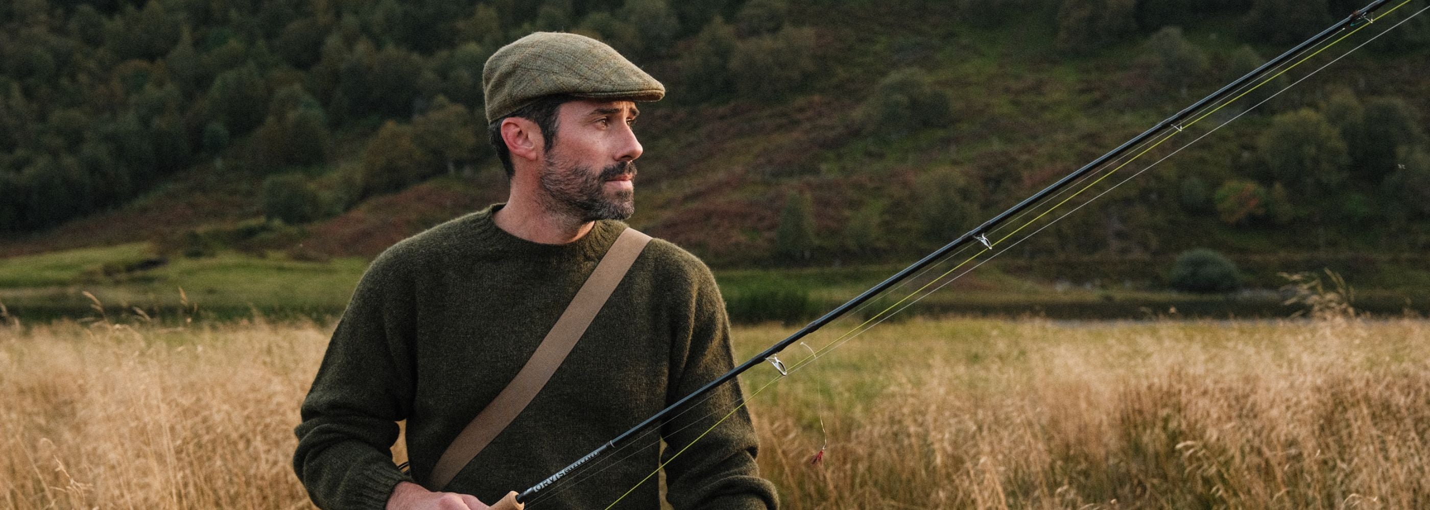 A man in a green sweater and flat cap stands in a grassy field, holding a fishing rod. Trees and hills are visible in the background under soft daylight.