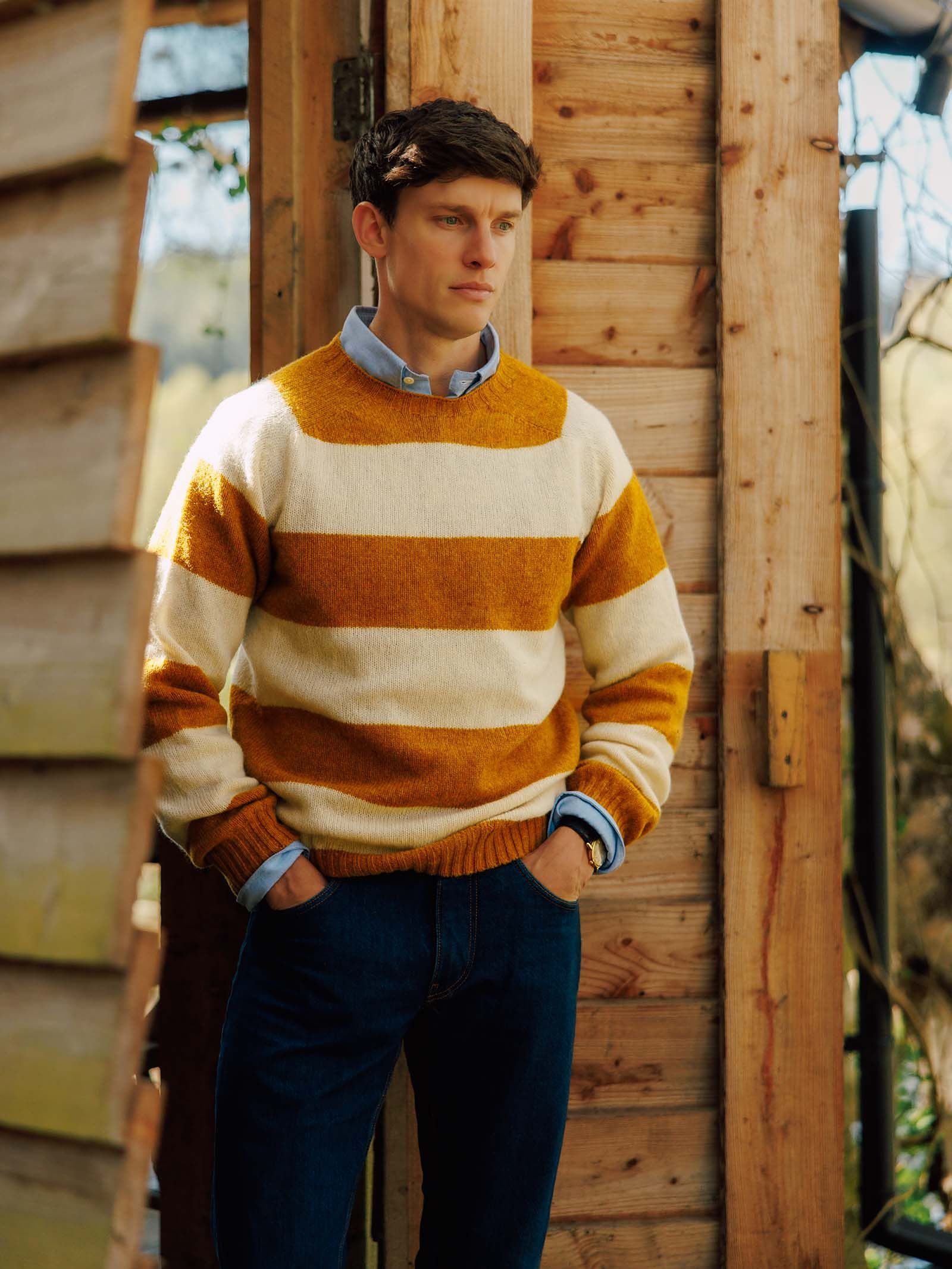 A man stands outside a wooden building, wearing the Campbell’s of Beauly Shetland Stripe Crew Jumper in yellow and white over a blue collared shirt and dark jeans, with his hands in his pockets.