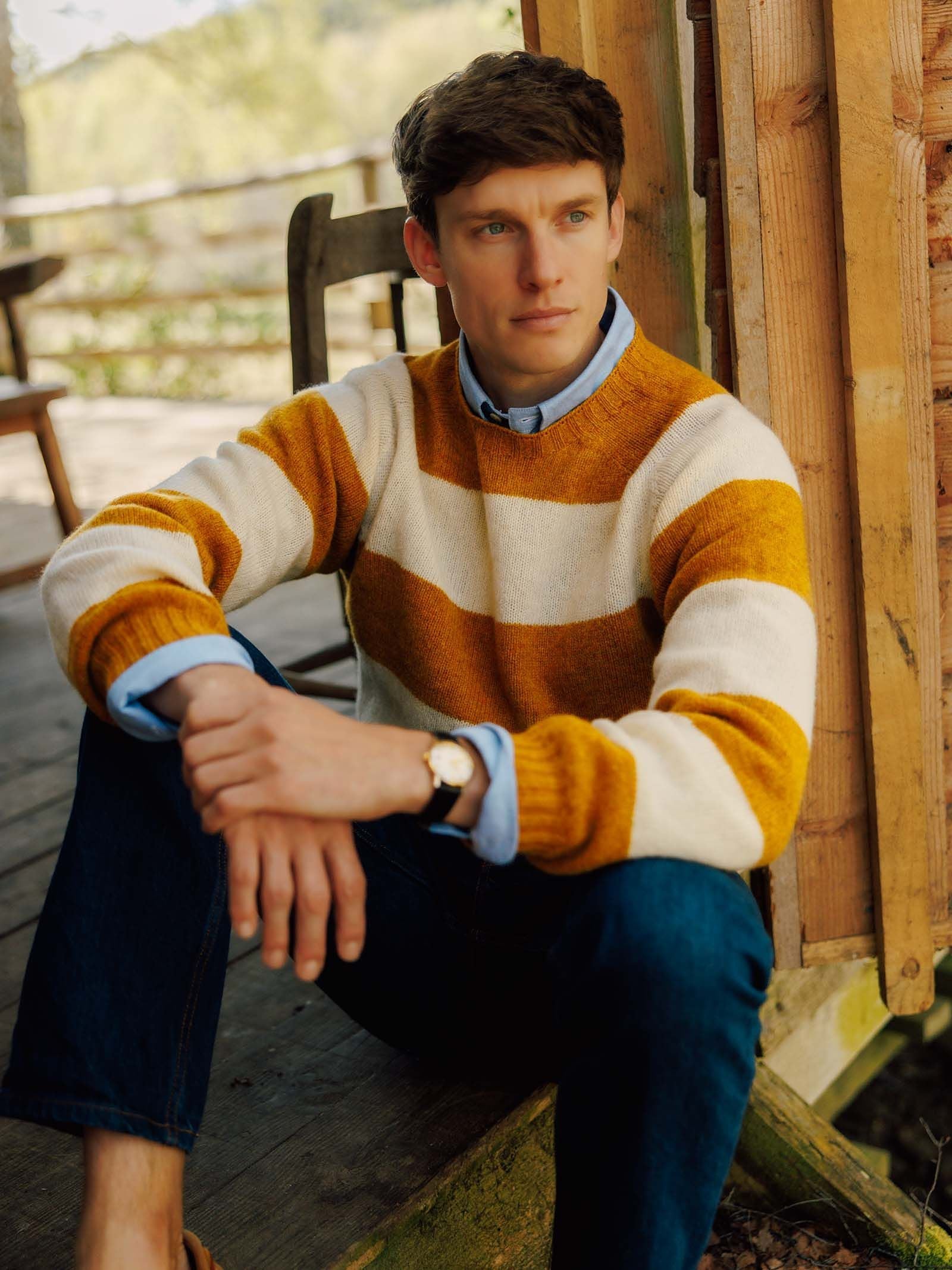 A man sits on a wooden porch, dressed in the Campbell’s of Beauly Shetland Stripe Crew Jumper—its yellow and white stripes layered over a blue collared shirt and jeans. Thoughtfully gazing to the side, he is surrounded by rustic wooden boards and natural light.