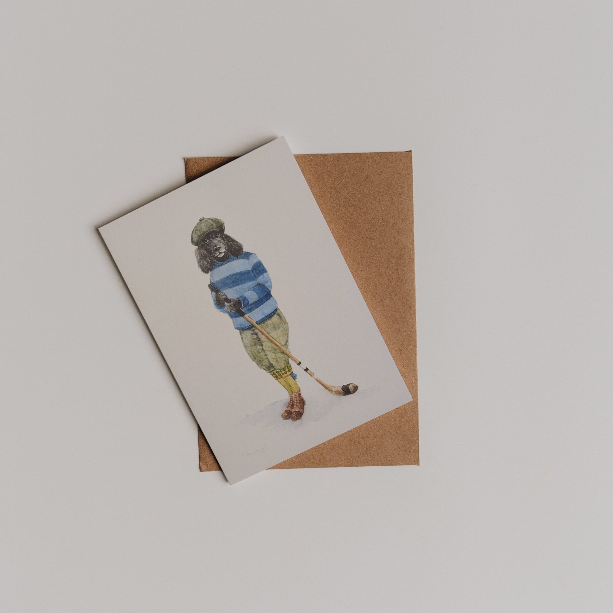 The Shinty Player Greeting Card by Campbell's of Beauly features an illustrated player in a cap, blue-striped sweater, and green pants holding a hockey stick, shown on a brown envelope. Designed in collaboration with Sophie Murray.