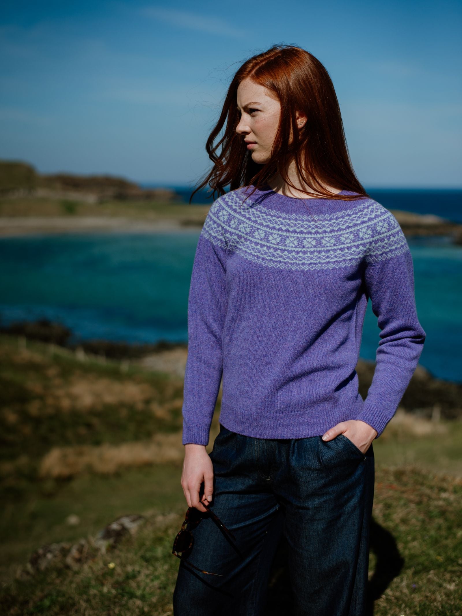 A woman with long red hair wears the Campbell’s of Beauly Auld Stock Two-Colour Fairisle Yoke Jumper and dark pants, standing by a turquoise sea and grassy landscape, holding sunglasses and looking to the side under a clear blue sky.
