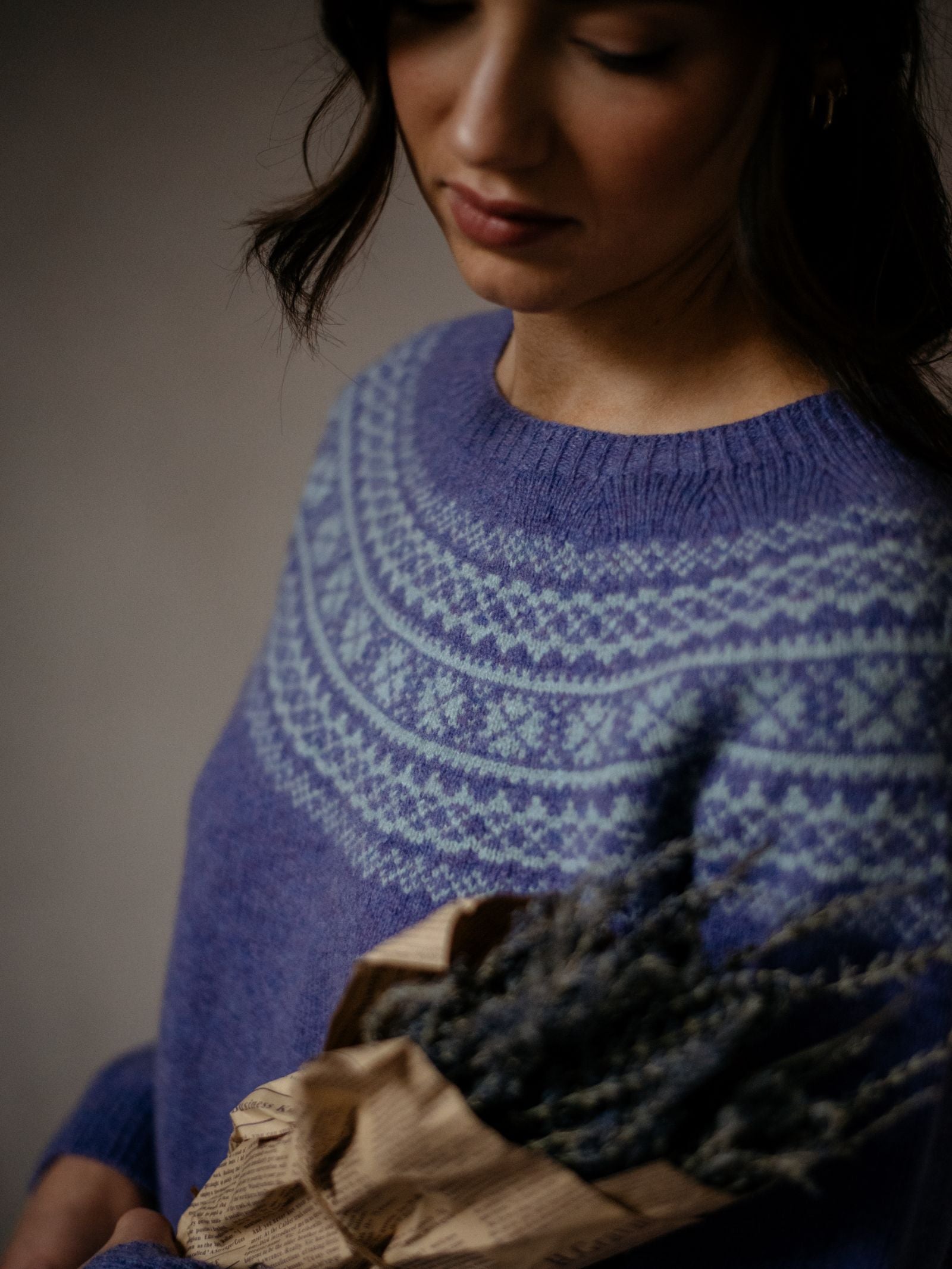 A woman wears the Campbell's of Beauly Auld Stock Two-Colour Fairisle Yoke Jumper, holding dried lavender wrapped in brown paper as she looks down softly under gentle, muted lighting.