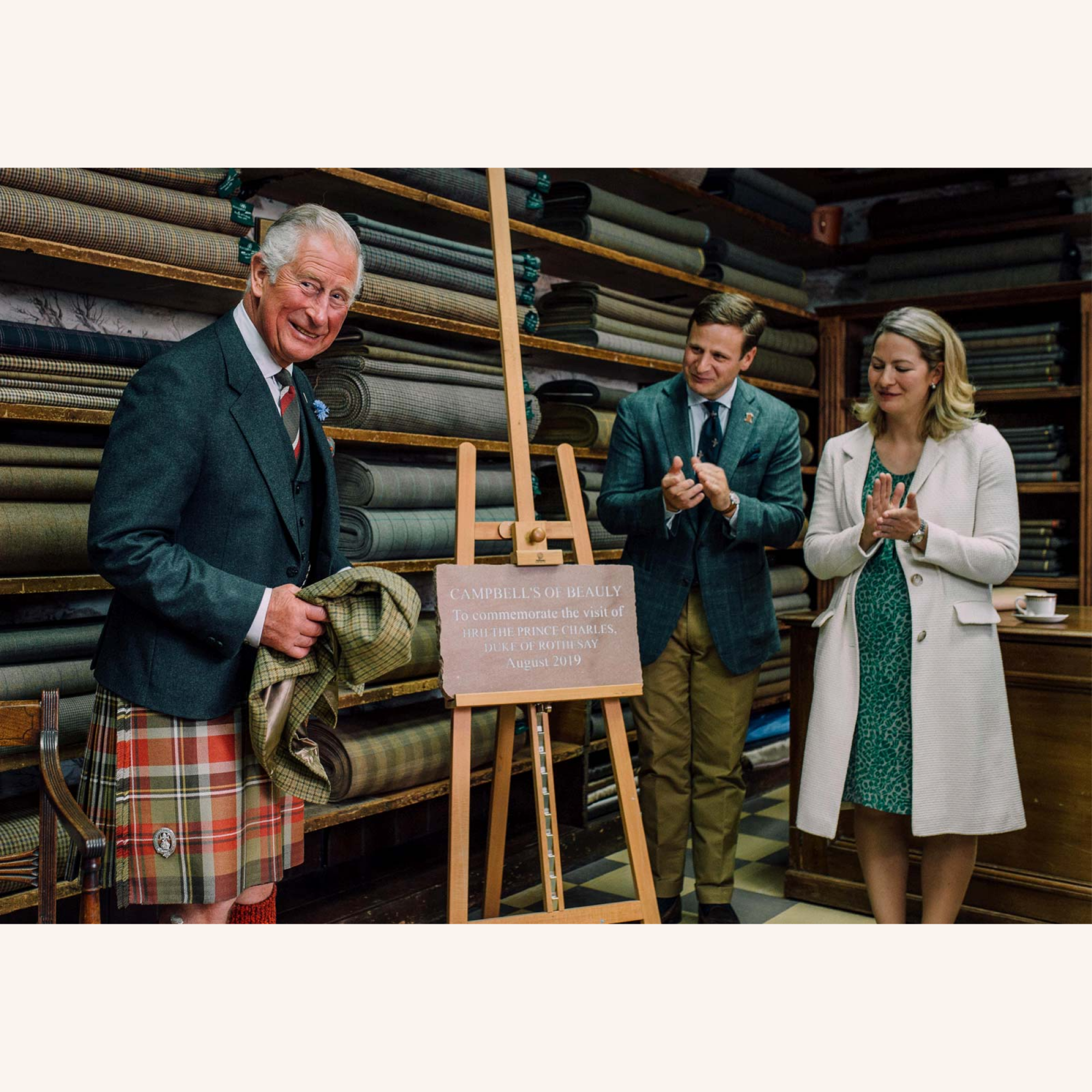 An older man in a plaid kilt unveils a commemorative plaque in a fabric shop as a man and woman beside him clap. Rolls of fabric are stacked on shelves behind them.