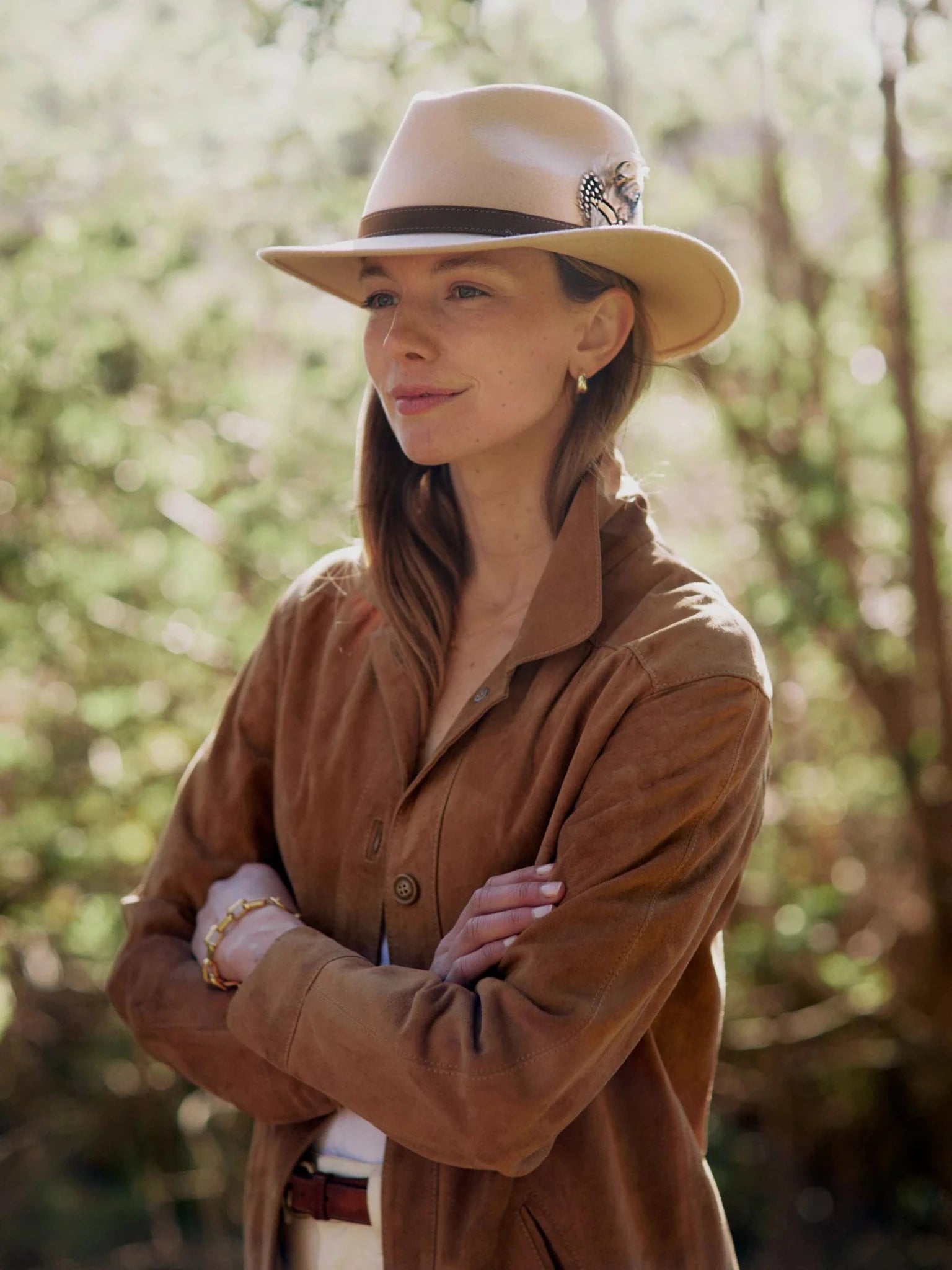 A woman wearing a tan suede jacket and a Hicks & Brown Auld Stock Gamebird Fedora with feather detail stands outdoors, arms crossed, surrounded by softly blurred greenery in natural light.