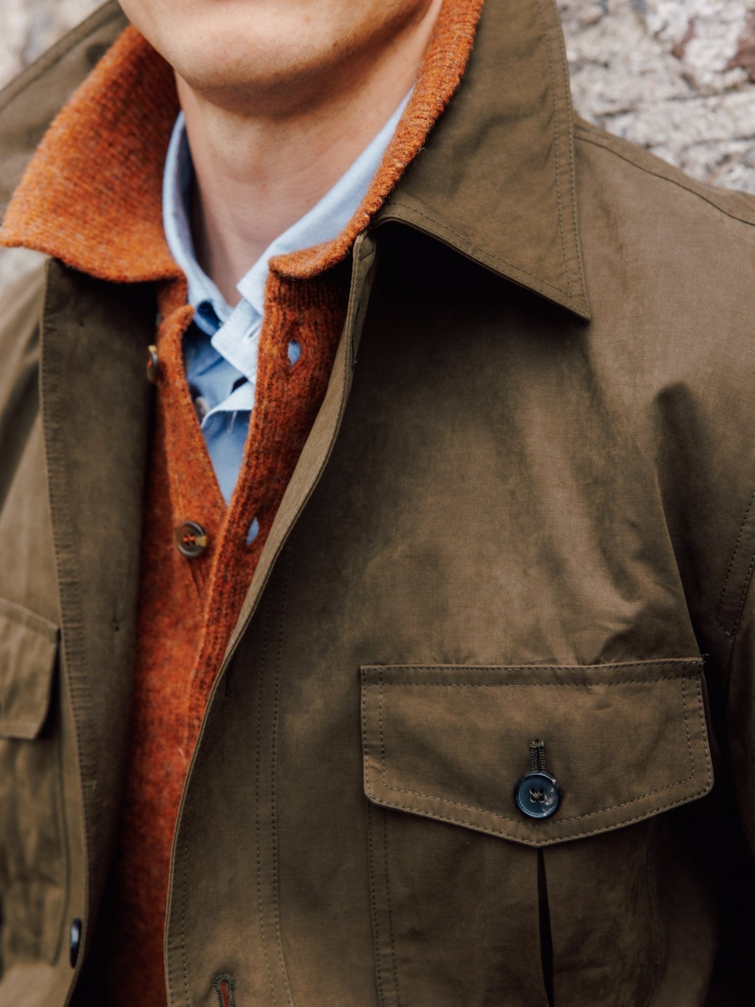 A close-up shows a person in the Campbell's of Beauly Aigas Field Jacket layered over an orange sweater and light blue collared shirt, standing by a stone wall with their face partially out of frame.