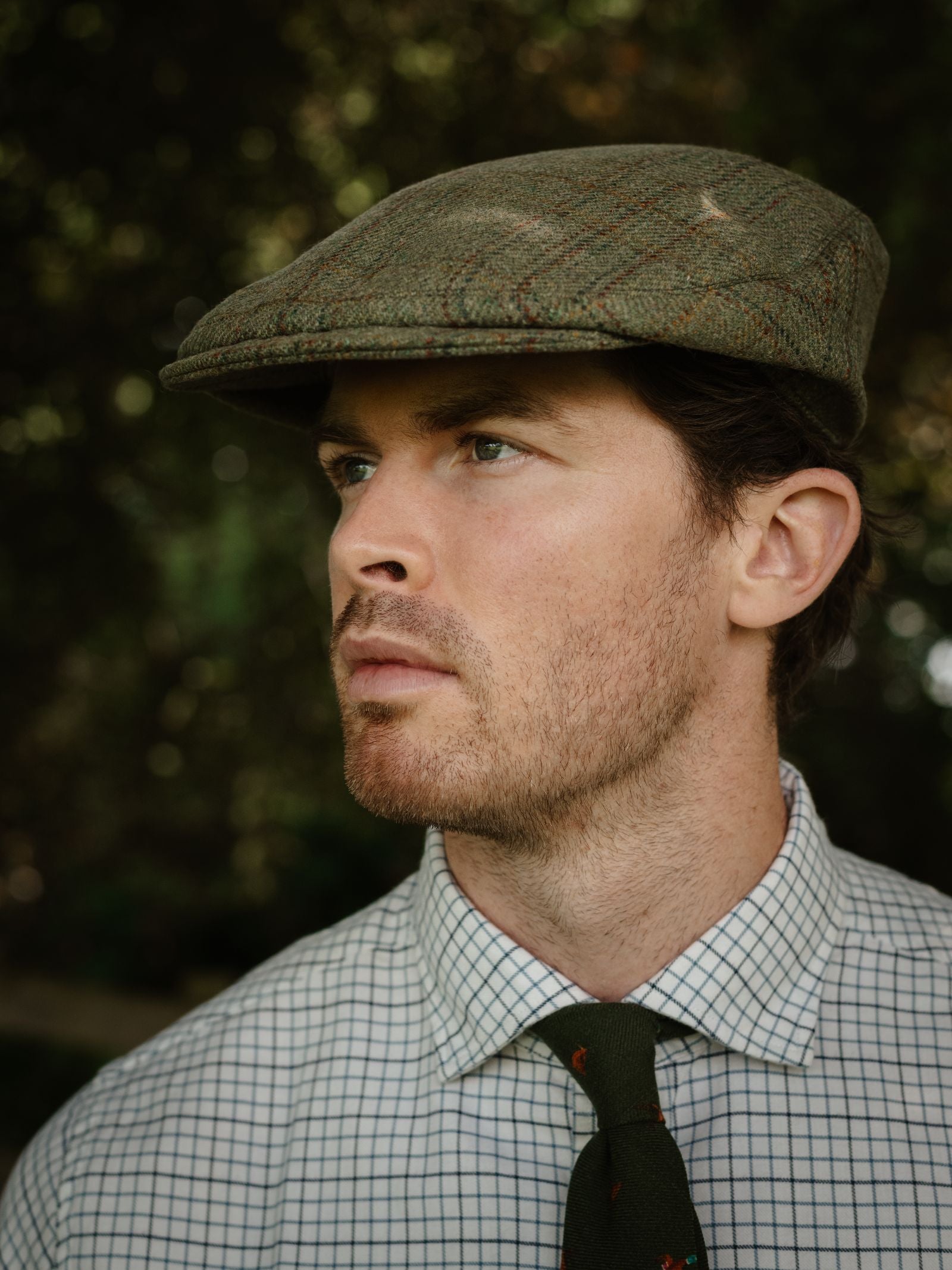 A man wears the Campbell's of Beauly Unisex Beauly Cap in green tweed, paired with a white checkered shirt and green tie, looking off to the side against a blurred outdoor background.