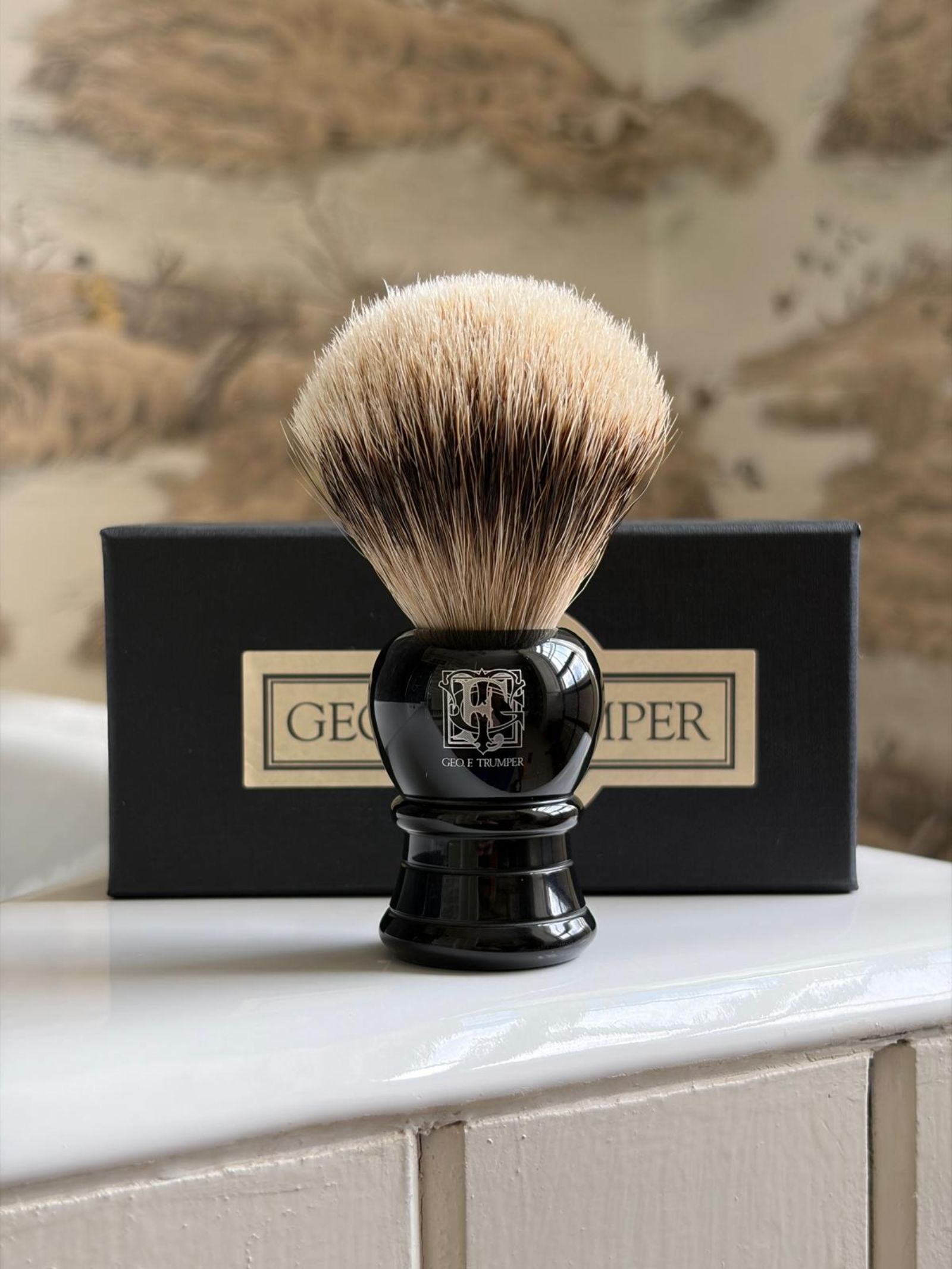 A Geo. F. Trumper Black Shaving Brush with dense bristles stands before its black box on a white bathroom countertop, promising a luxurious lather against the blurred patterned wall in the background.