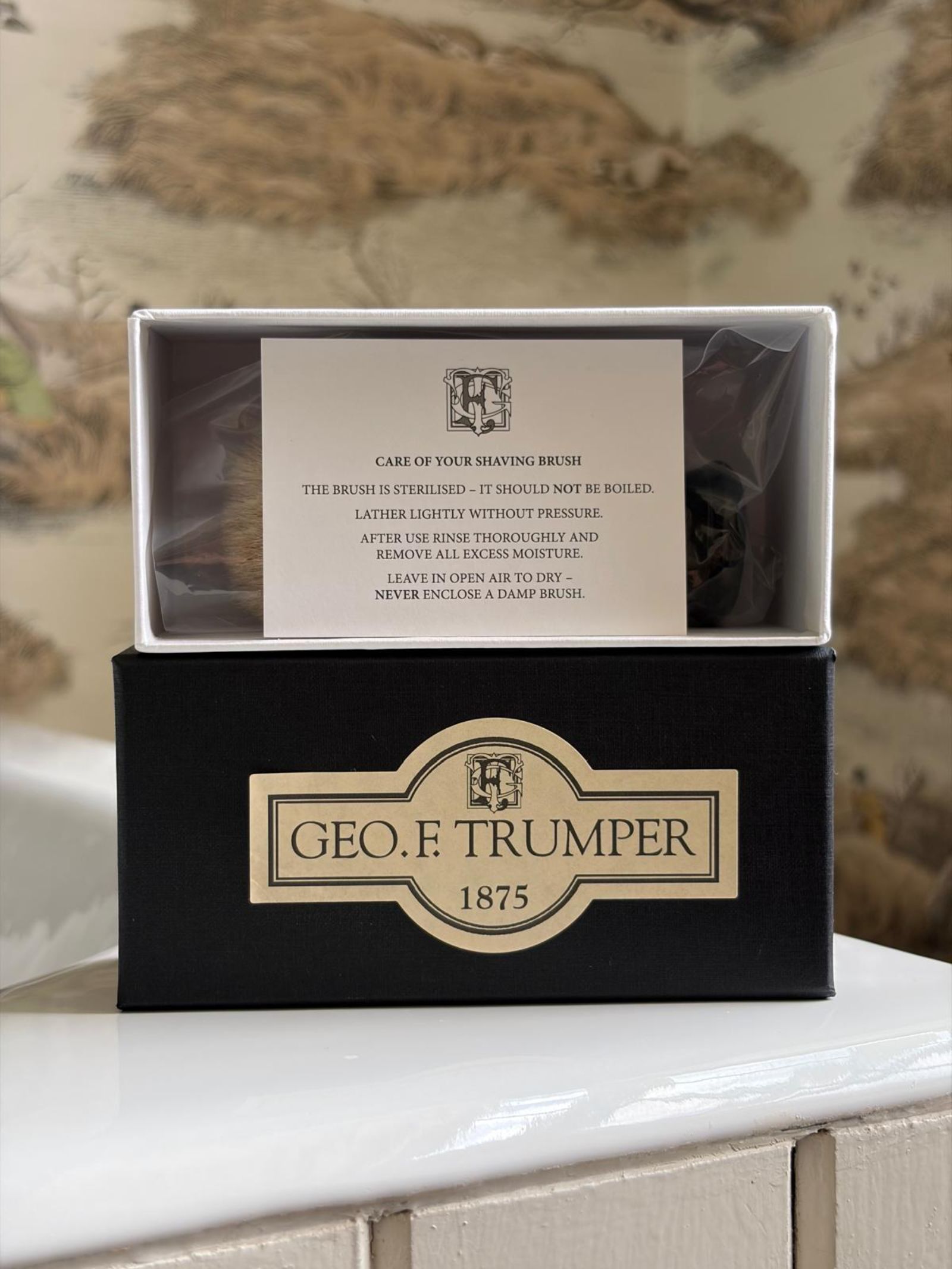 A Geo. F. Trumper Black Shaving Brush box sits open on a bathroom surface, revealing the care card inside; the elegant black box with gold branding ensures a comfortable shave and luxurious lather.