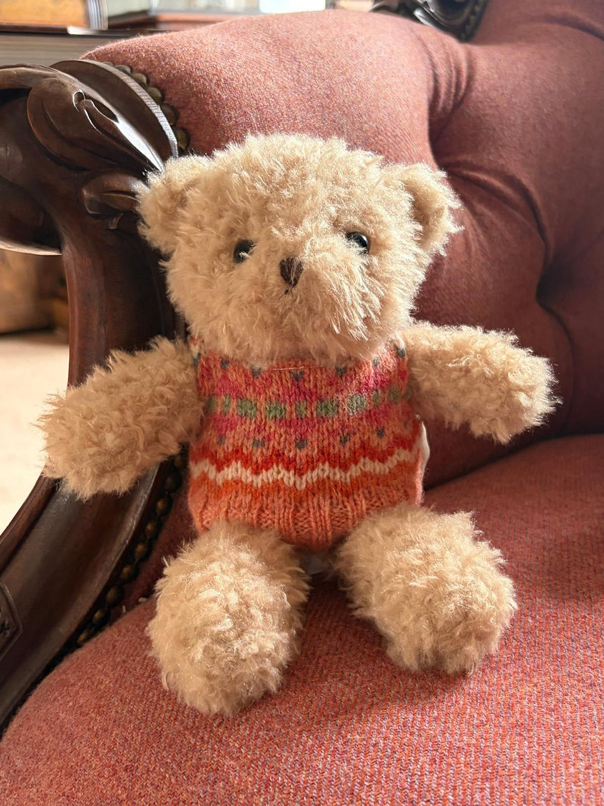 Bobby the Bear, a small and fluffy teddy from Campbell’s of Beauly, sits on an upholstered reddish armchair with wooden trim, wearing a red and orange knitwear-inspired sweater.