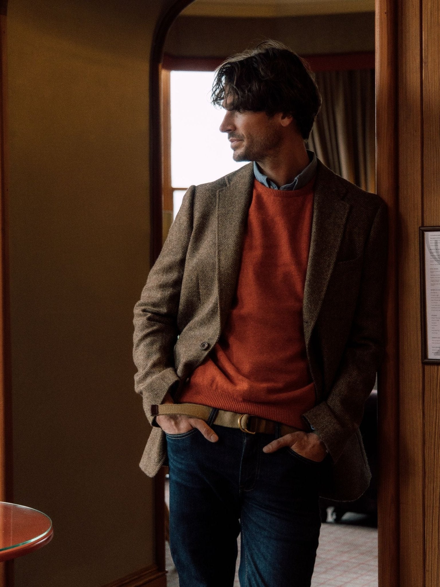 A man with medium-length dark hair stands indoors, wearing the Campbell's of Beauly Campbell's House Jacket in Brown Herringbone over an orange sweater and blue jeans, hands in his pockets as he looks out a window.