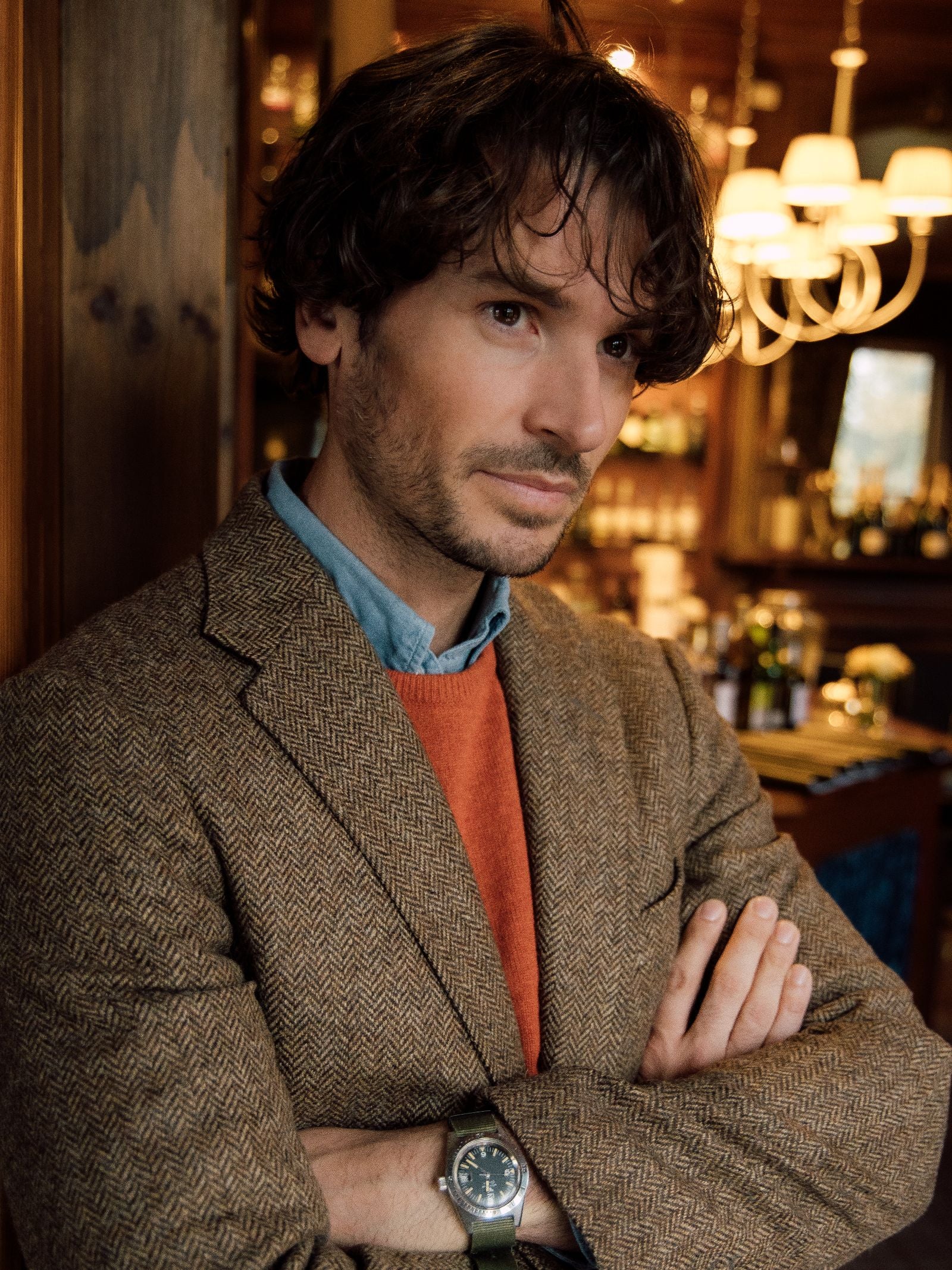 A man with wavy brown hair, wearing the Campbell's of Beauly Campbell's House Jacket in brown tweed over an orange sweater and blue shirt, stands indoors with arms crossed amid warm lighting and a chandelier.
