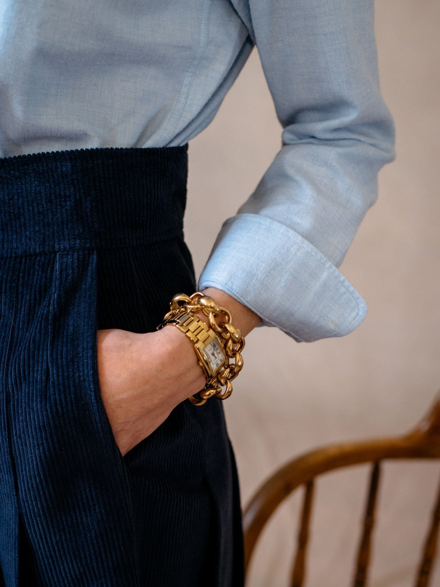 A person wears the Campbell's of Beauly Auld Stock Brushed Cotton Shirt Blue with dark blue corduroy pants, one hand in their pocket, showing a chunky gold watch and bracelet. A wooden chair is partly visible in the background.
