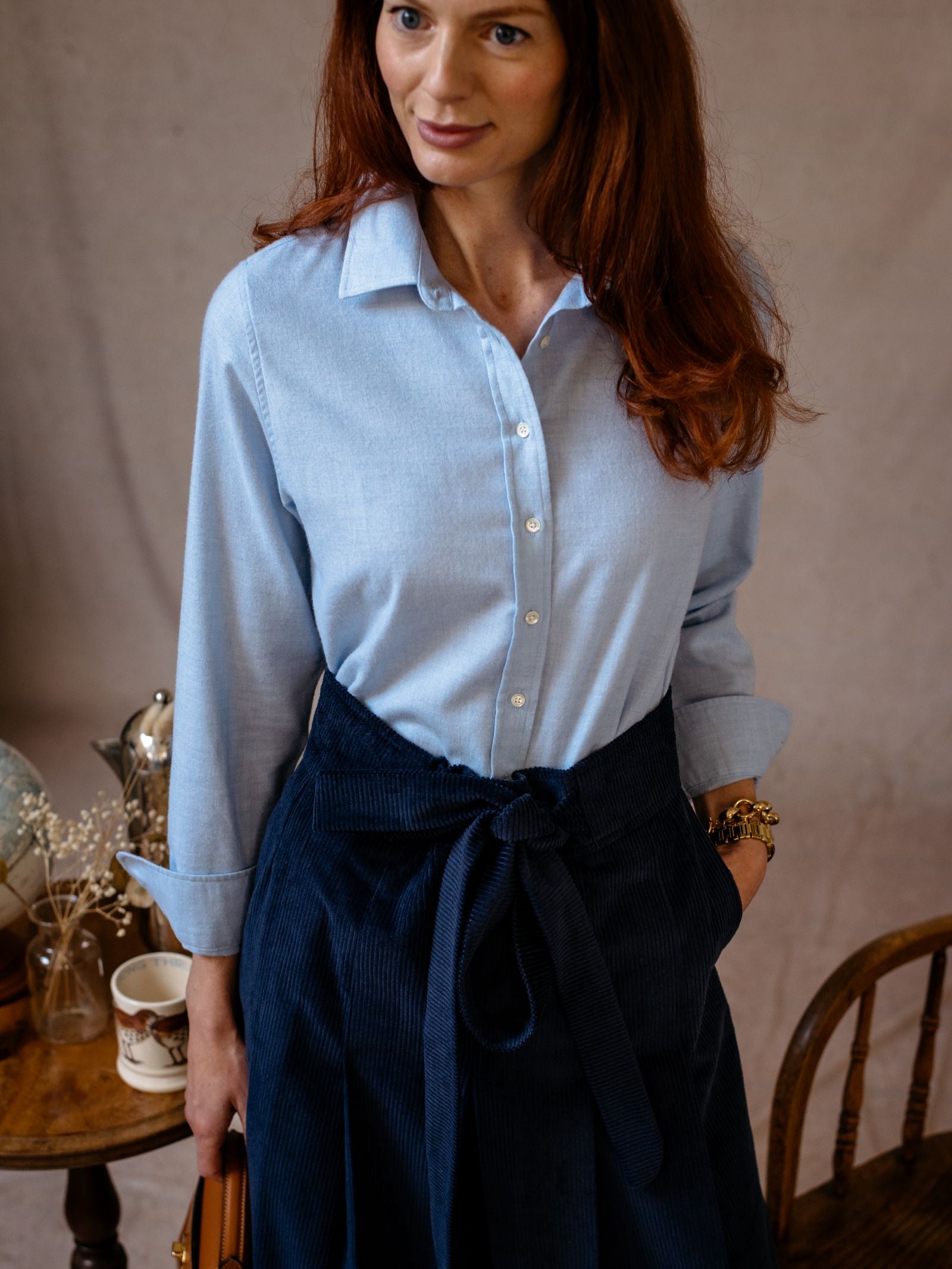 A woman with long reddish-brown hair wears the Auld Stock Brushed Cotton Shirt Blue by Campbell's of Beauly, tucked into a dark blue high-waisted skirt, standing indoors near a wooden chair and a small table with a cup and dried flowers.